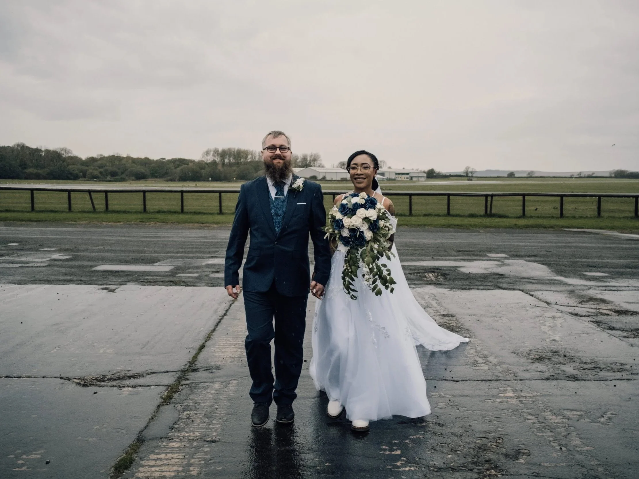 A newlywed couple walking hand-in-hand outdoors on a cloudy day, with the bride holding a bouquet of white and blue flowers. The groom has glasses, a beard, and is dressed in a dark suit, while the bride is wearing a white wedding dress and white boots.