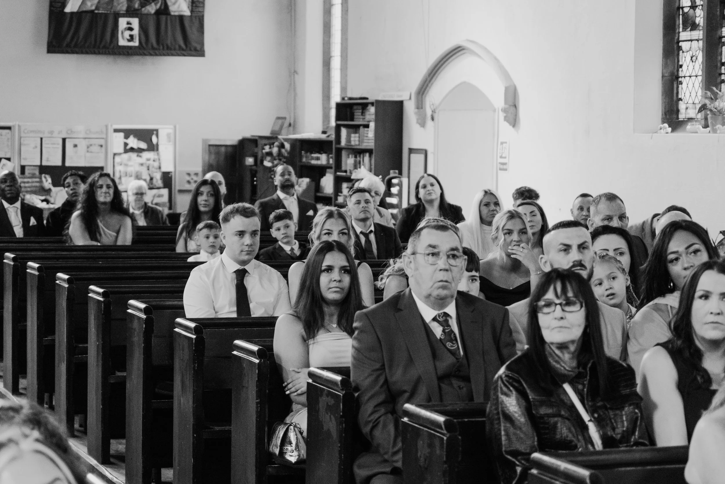 People seated inside a church during a service or ceremony, with various age groups and diverse appearances, facing forward.