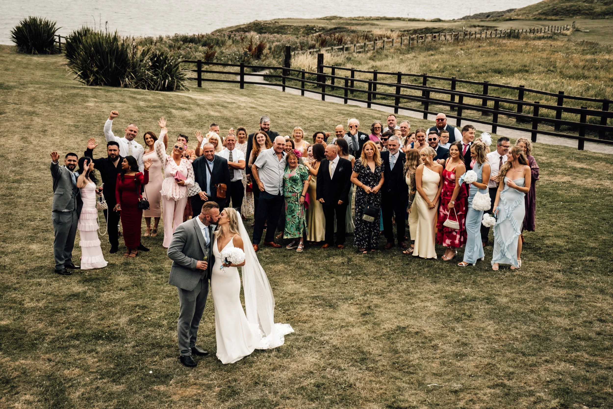 A large group of people gathered outdoors for a wedding celebration on a grassy area near the coast, with some waving and smiling. The bride and groom are in the foreground, with the bride in a white gown and the groom in a gray suit, sharing a kiss.