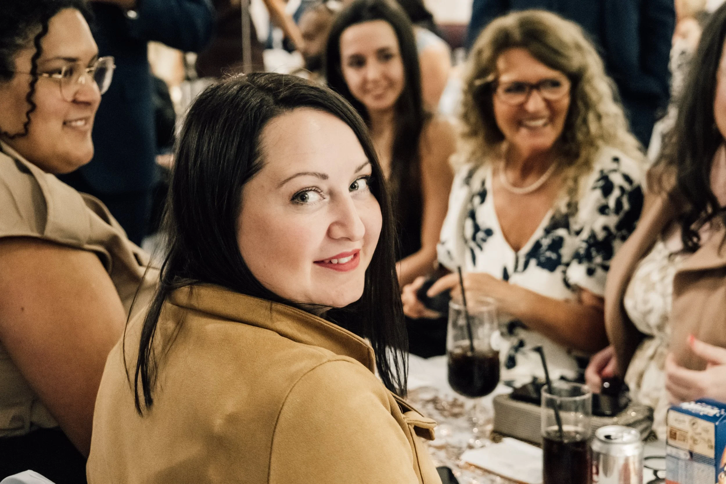 A group of women sitting around a table, socializing at a gathering or event, with a focus on a woman with dark hair and a tan blazer in the foreground.