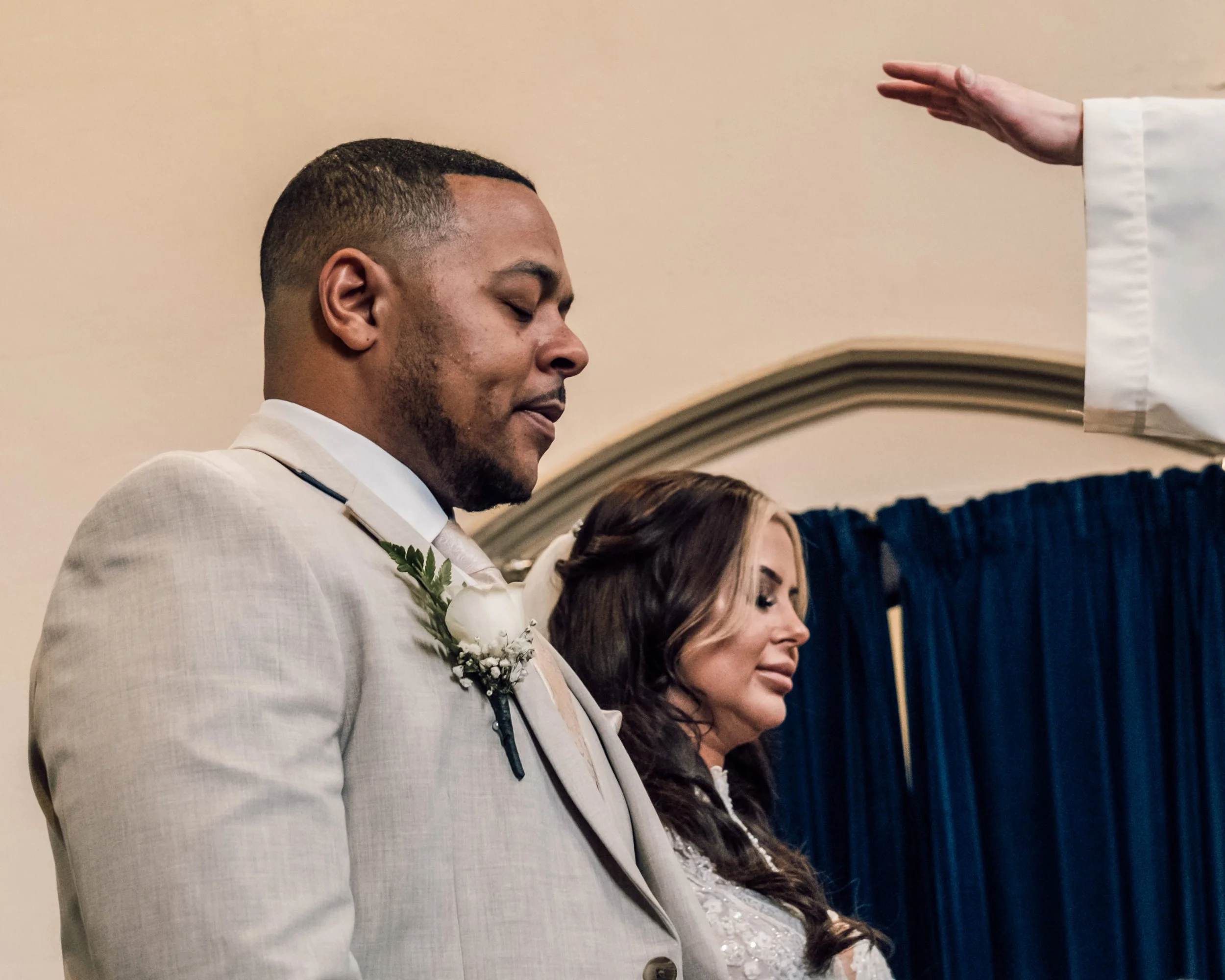 A man with short hair and a beard, dressed in a light-colored suit, appears to be praying during a ceremony. A woman is next to him, also with her eyes closed in prayer. An adult hand with a white sleeve extends horizontally over them, possibly perfo