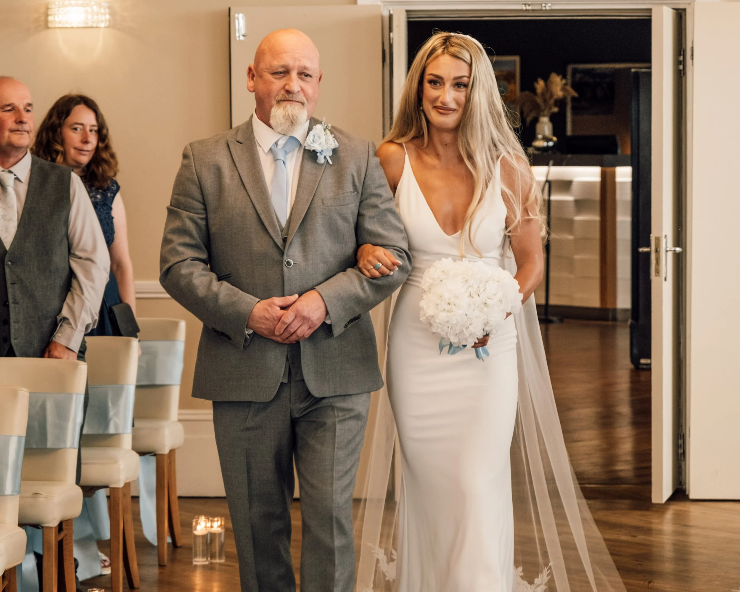 A bride walking arm-in-arm with an older man, possibly her father, during a wedding ceremony. She is holding a bouquet of white flowers and wearing a white wedding dress. The man is dressed in a gray suit with a white shirt and a light blue tie, and 