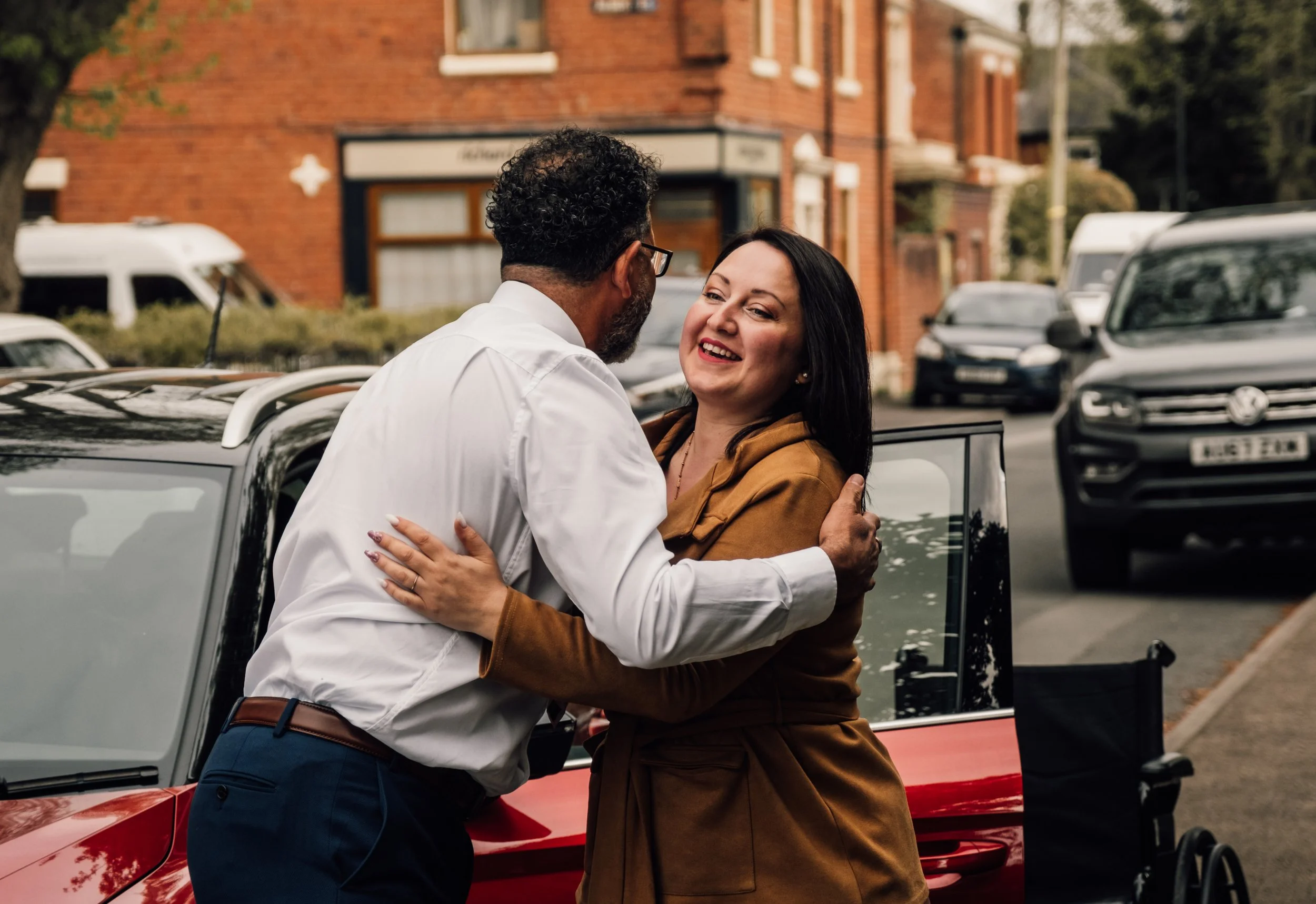 A man and a woman embrace and smile at each other on a street with parked cars and a brick building in the background.