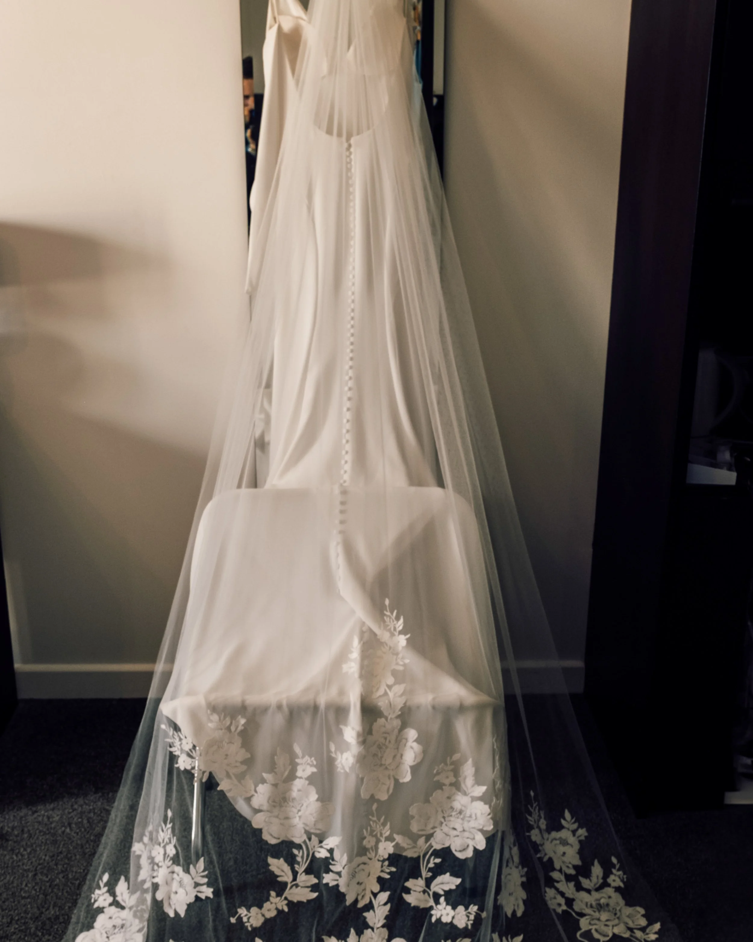 A wedding dress with floral embroidery on the bottom, hanging behind a sheer veil on a dress form.