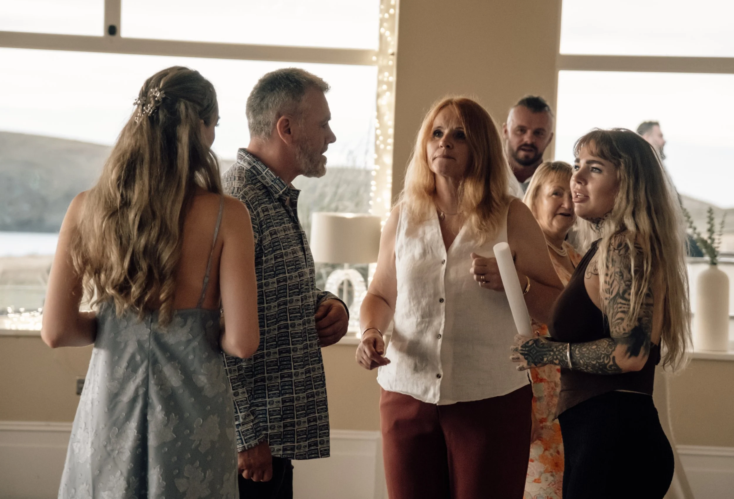 A group of people, mostly women, standing indoors, engaged in conversation at a social gathering, with large windows and natural light in the background.
