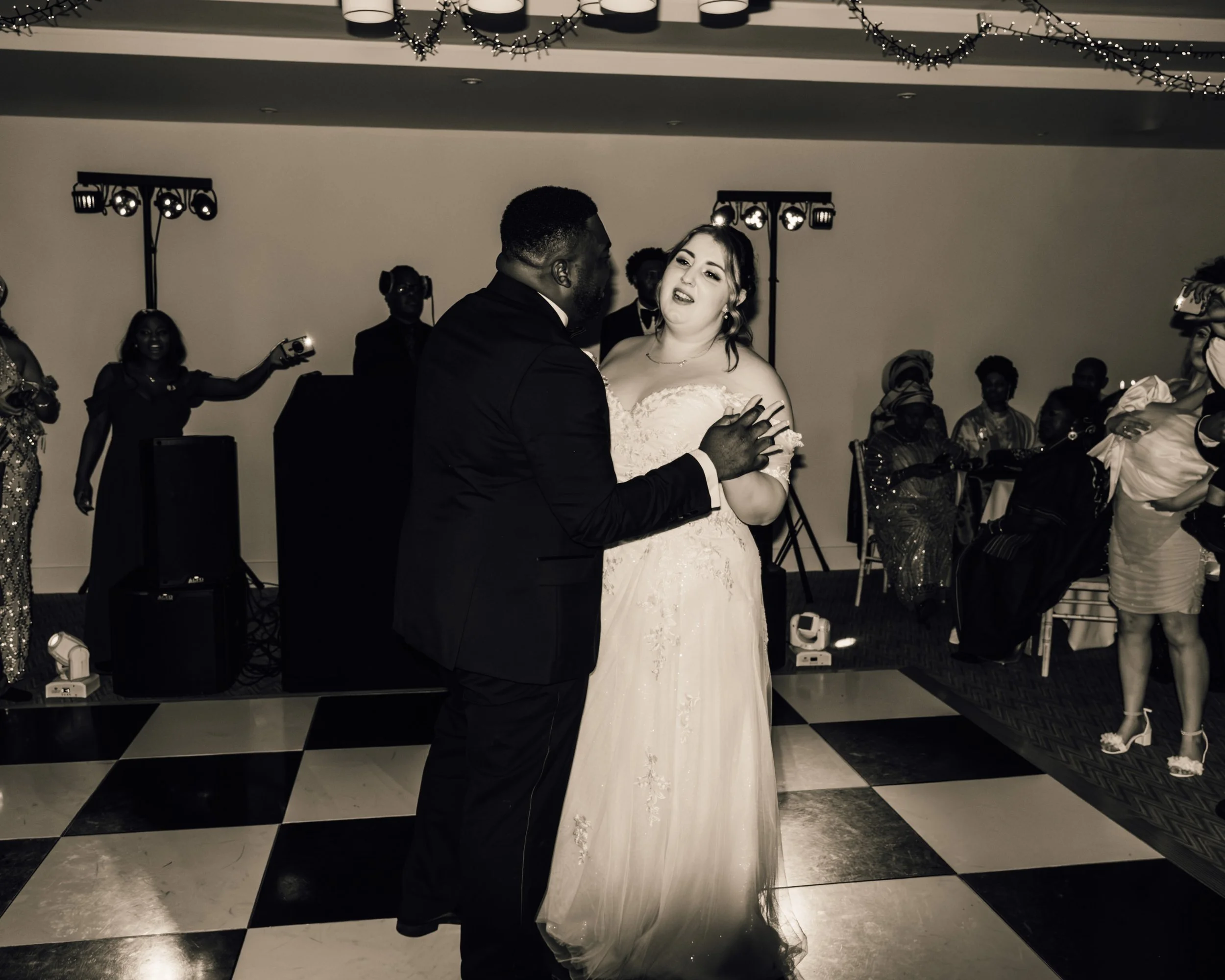 A bride and groom dancing at their wedding reception, with guests and photographers in the background. The bride wears a white wedding dress and the groom is in a dark suit. The venue has a black and white checkered floor and festive string lights ov