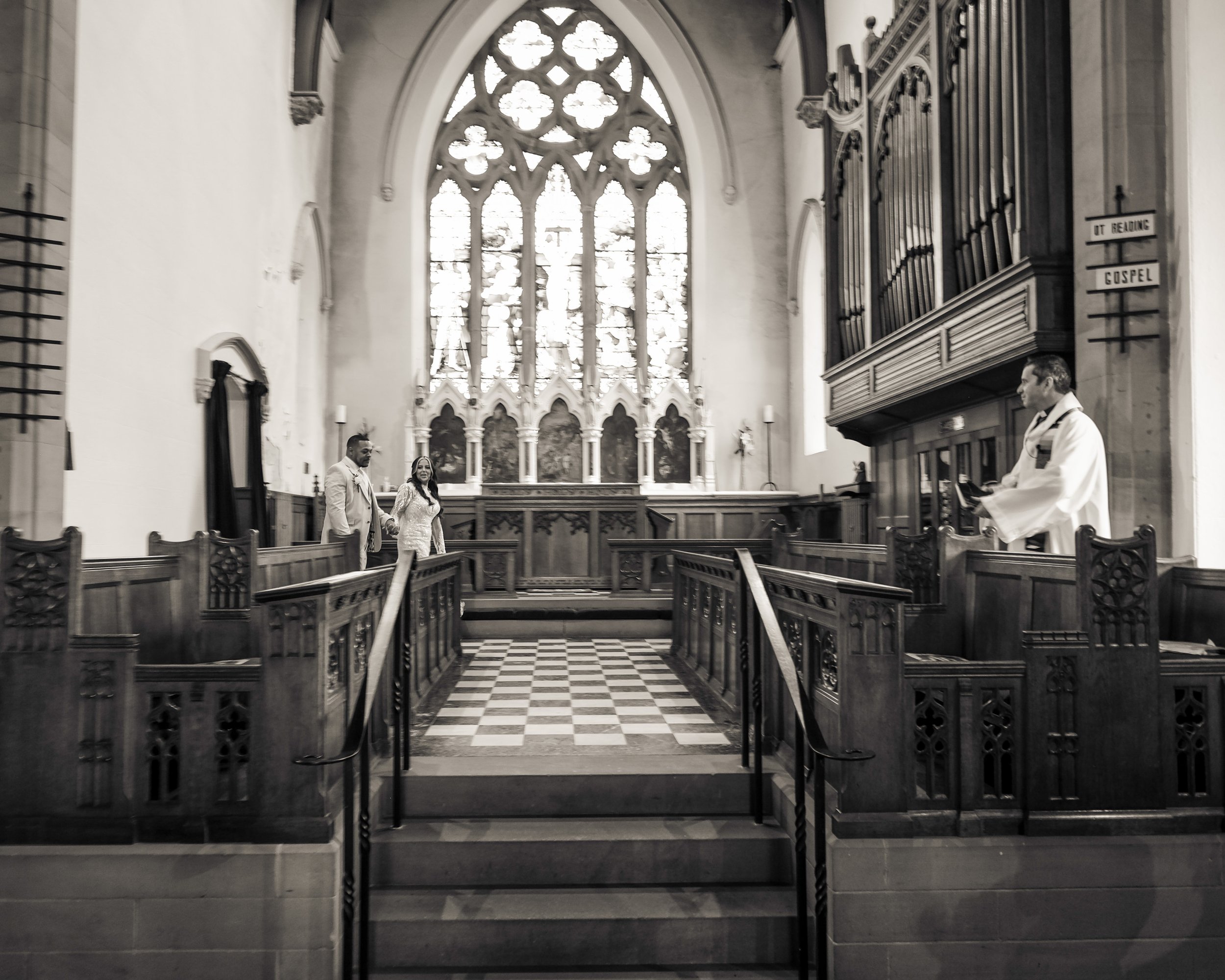 Interior of a church with stained glass windows, wooden pews, and a checkered stone floor. A man and woman are walking arm in arm at the front, with a person in white clergy attire on the right side near an organ. The church has tall windows and deco