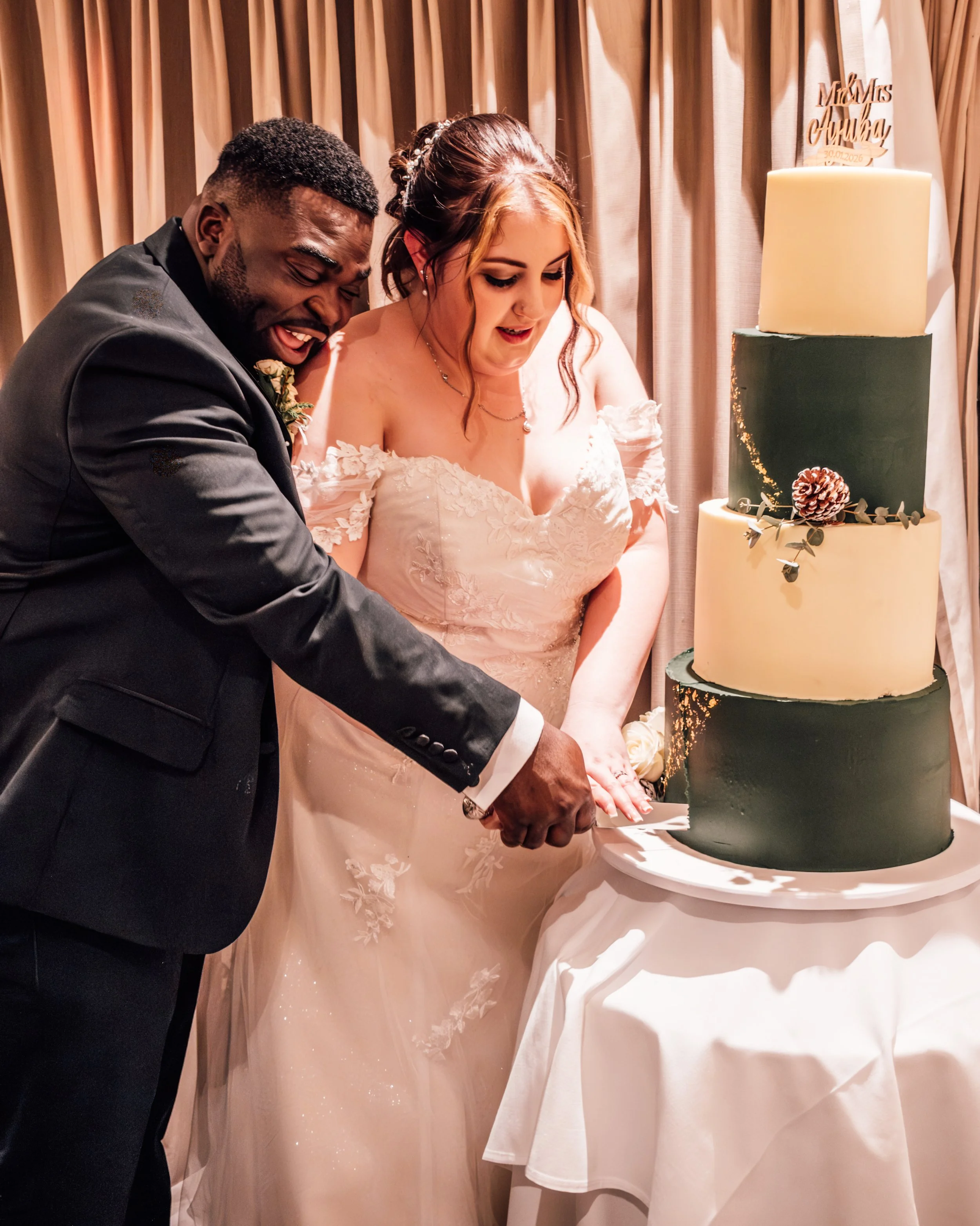 A bride and groom cut a wedding cake together. The bride wears a white off-shoulder wedding gown with lace details, and the groom wears a dark suit. On the right, there is a three-tiered wedding cake with green and cream colors, decorated with a pine
