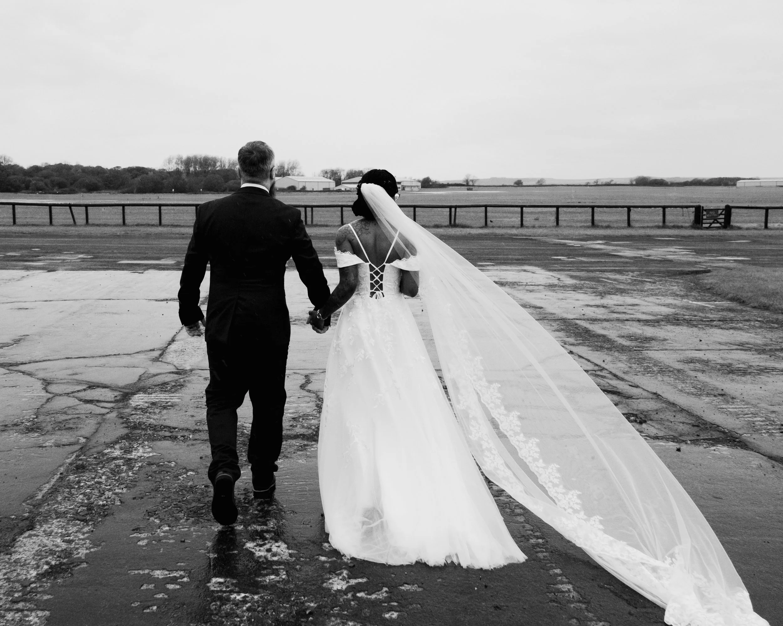 A black and white photo of a man and a woman, dressed in wedding attire, walking hand in hand on a tarmac runway, with an open field and some buildings in the background. The woman is wearing a wedding dress and veil, while the man is wearing a suit.