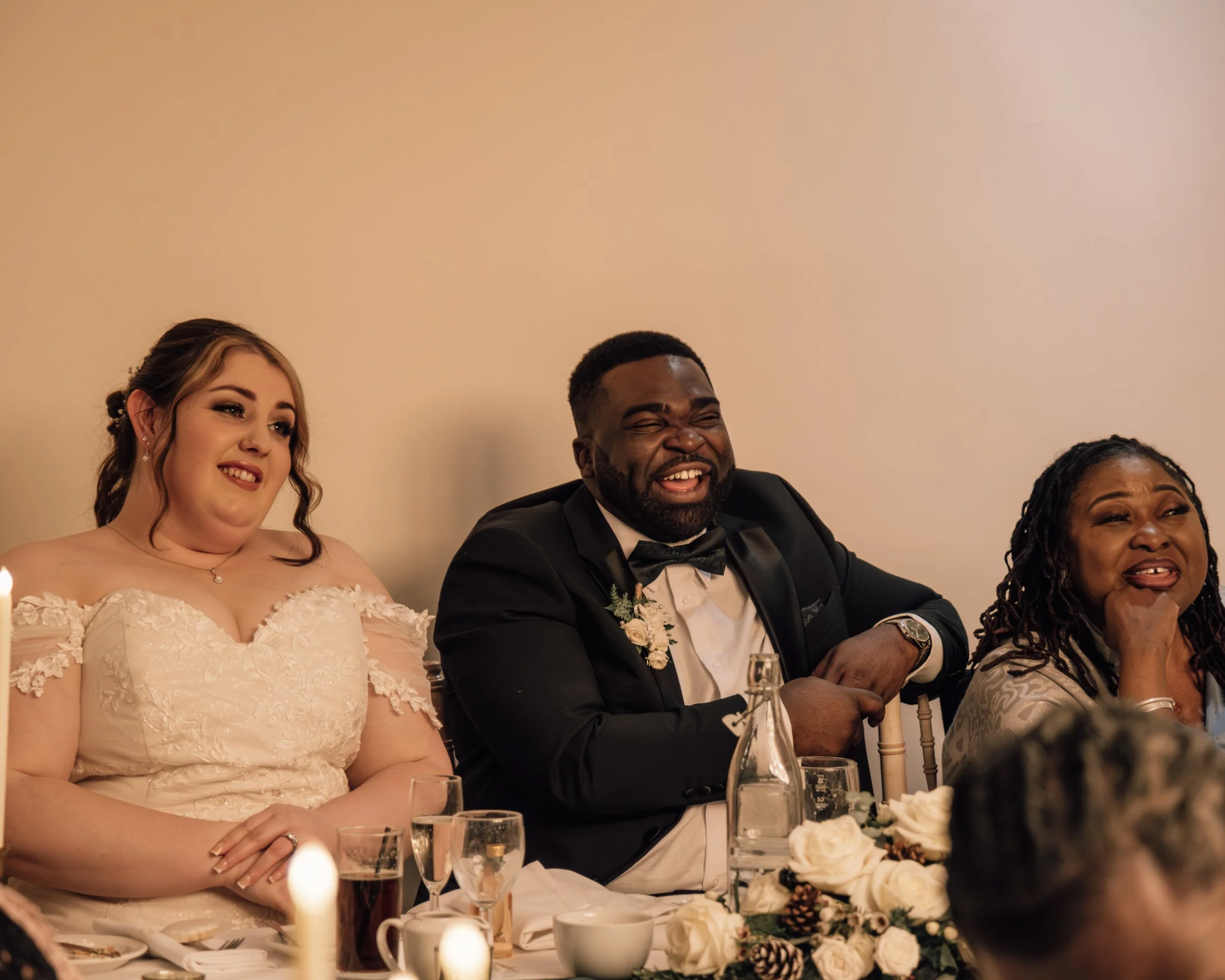 People sitting at a wedding reception table, smiling and enjoying the celebration. The table is decorated with flowers, pinecones, and candles.