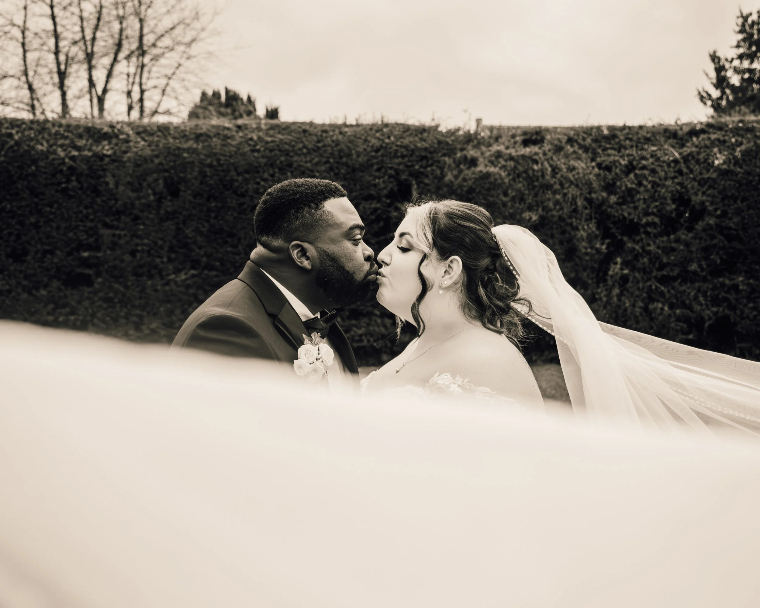 A black and white photo of a bride and groom about to kiss outdoors, with trees and a hedge in the background.