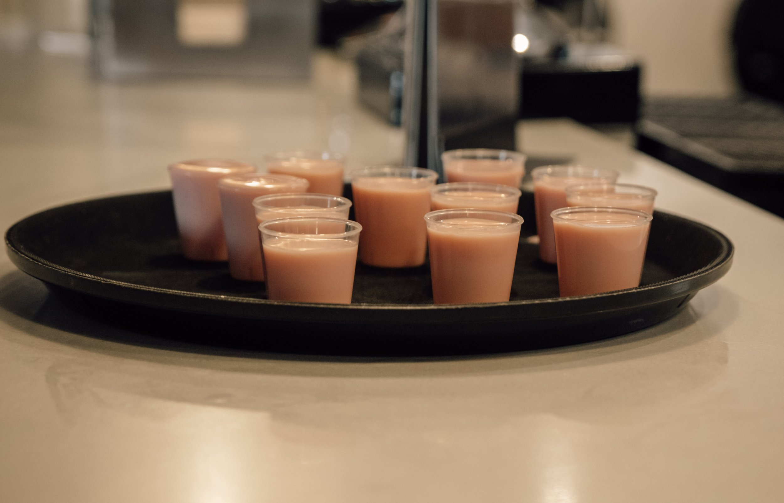 A black tray with multiple small plastic cups filled with a light brown liquid, likely a beverage, placed on a white surface.