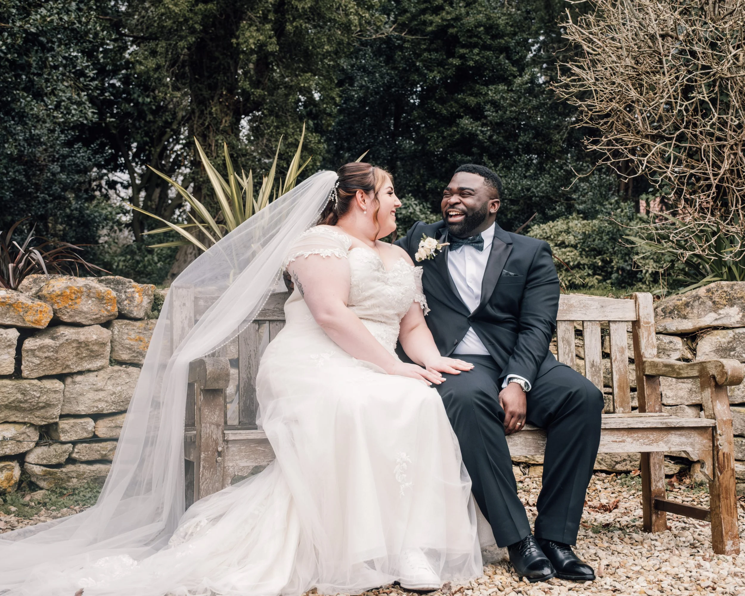 A bride and groom sitting on a wooden bench outdoors, smiling and looking at each other. The bride is wearing a white wedding dress with a long veil, and the groom is in a black tuxedo with a bow tie. They are surrounded by greenery and stone element