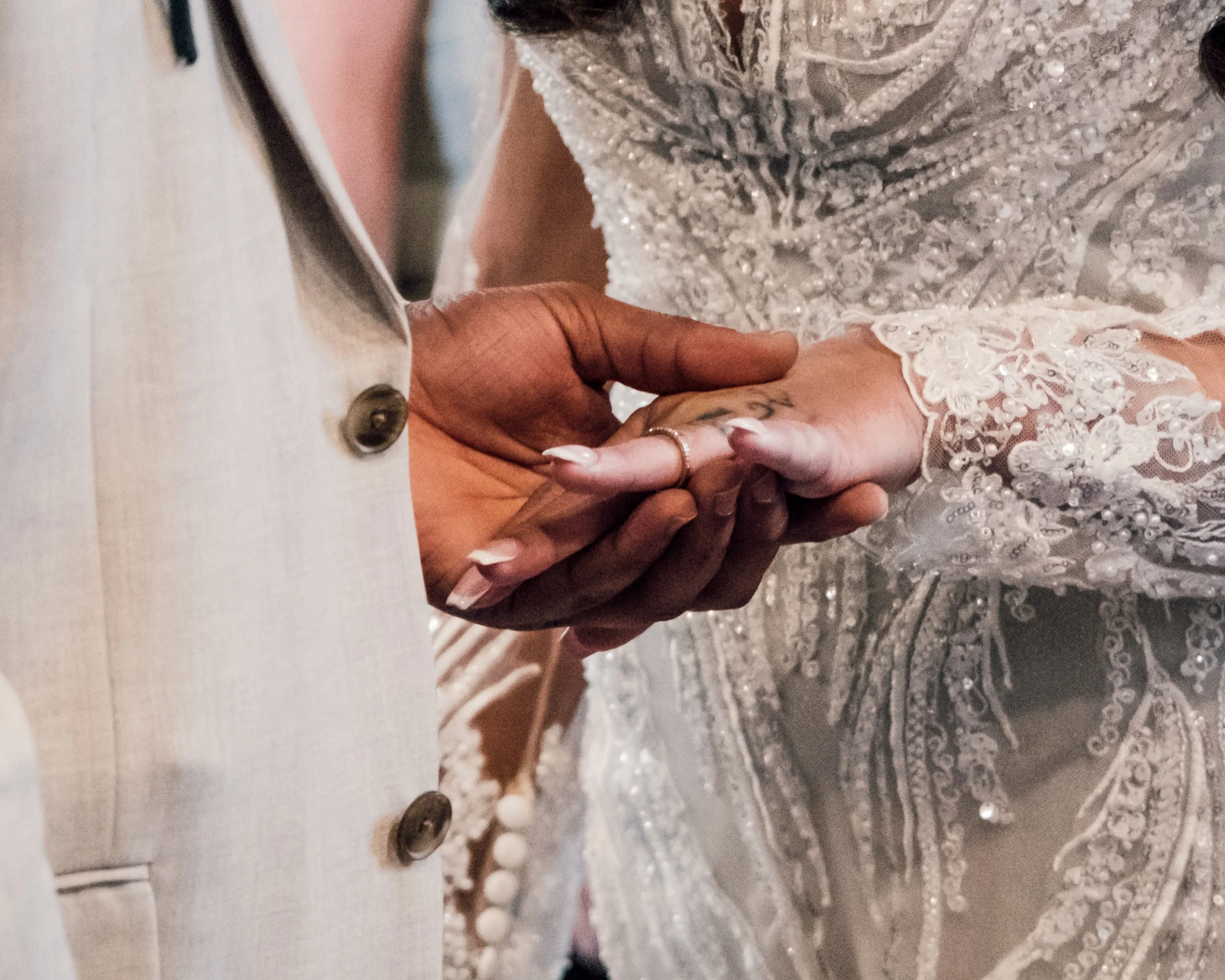 Close-up image of two hands holding each other, one hand is adorned with a ring and the backdrop features ornate, sequined, and embroidered fabric, likely a wedding dress.