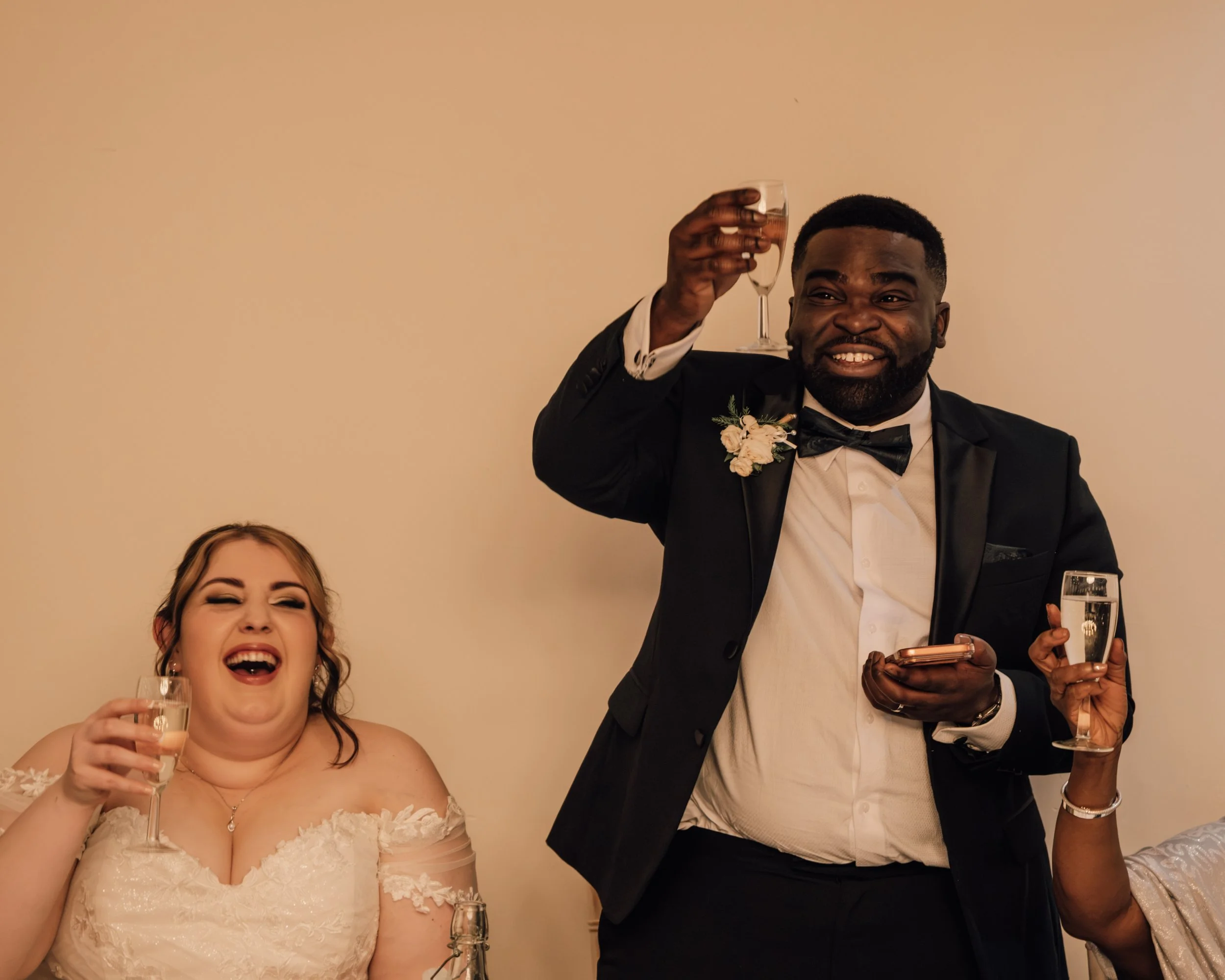 A joyful bride and groom celebrating at a wedding reception. The bride, with dark hair styled in an updo, is wearing a white lace wedding dress and holding a glass of champagne, laughing with her mouth open. The groom, a dark-haired man with a beard,