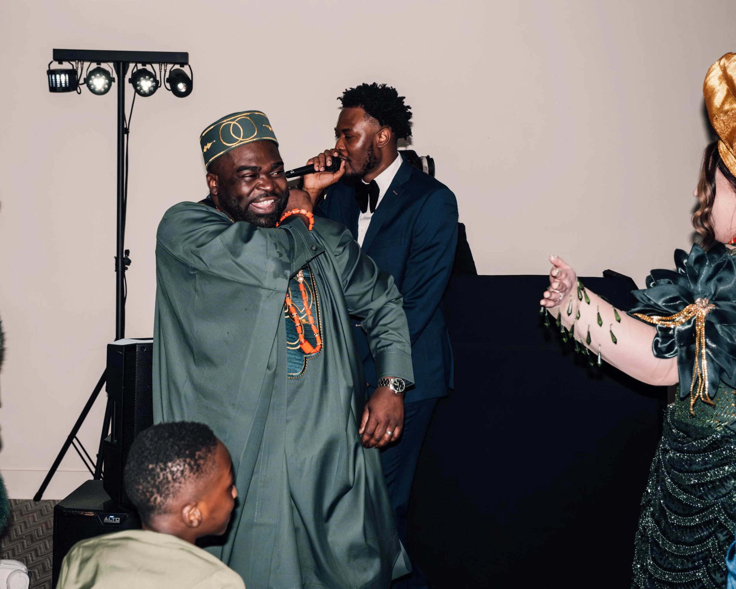 A man in traditional African attire and a woman in a green dress with gold accents dancing and singing with a microphone at an indoor event, with a DJ in the background.