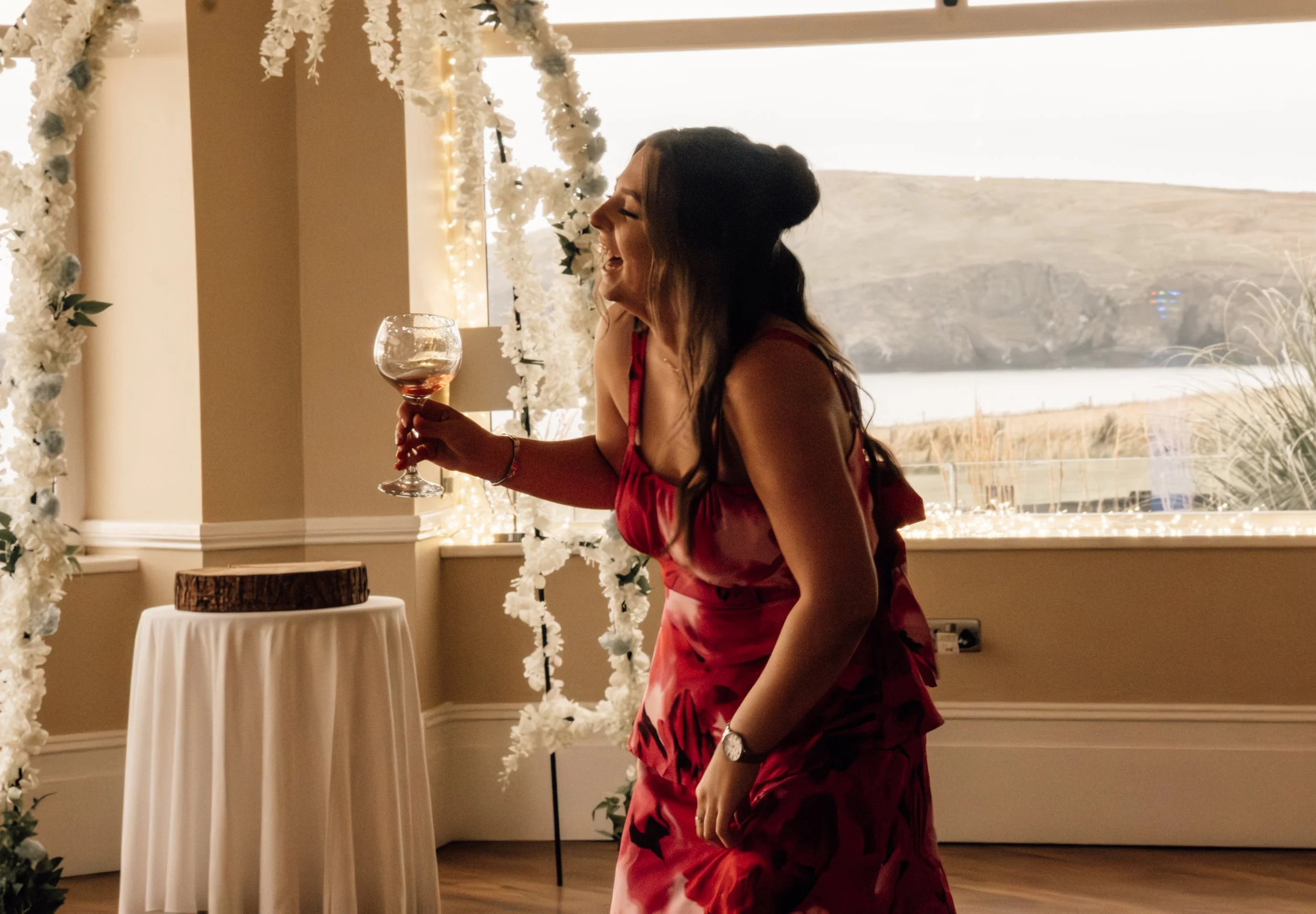 A woman in a red dress laughing and holding a glass of wine at an indoor event, decorated with white floral arrangements, with a window showing an outdoor landscape in the background.