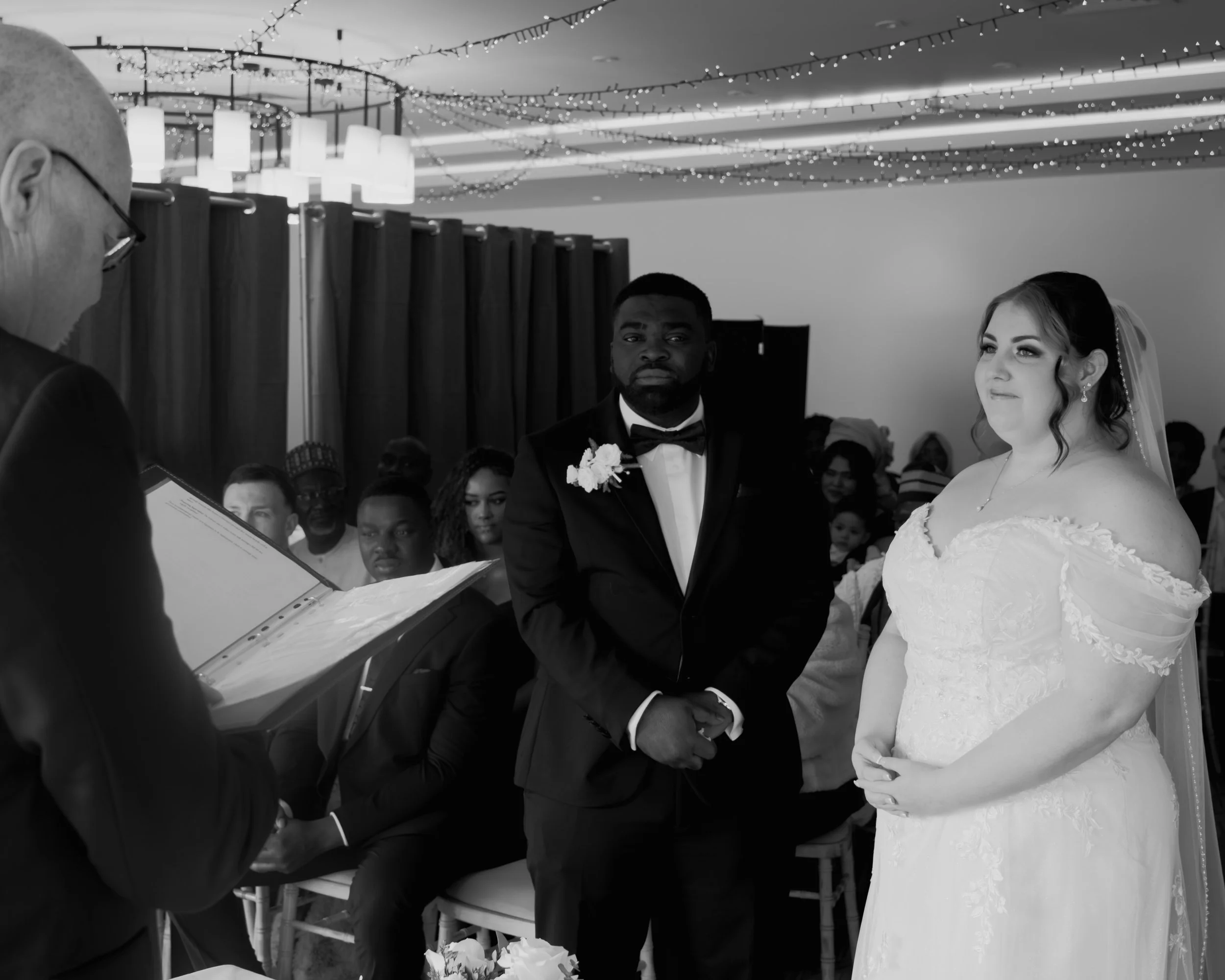 A wedding ceremony with a bride and groom standing together, holding hands, in front of an officiant. The groom is wearing a tuxedo with a bow tie and a flower boutonniere, and the bride is in a lace wedding dress with a veil. There are guests seated
