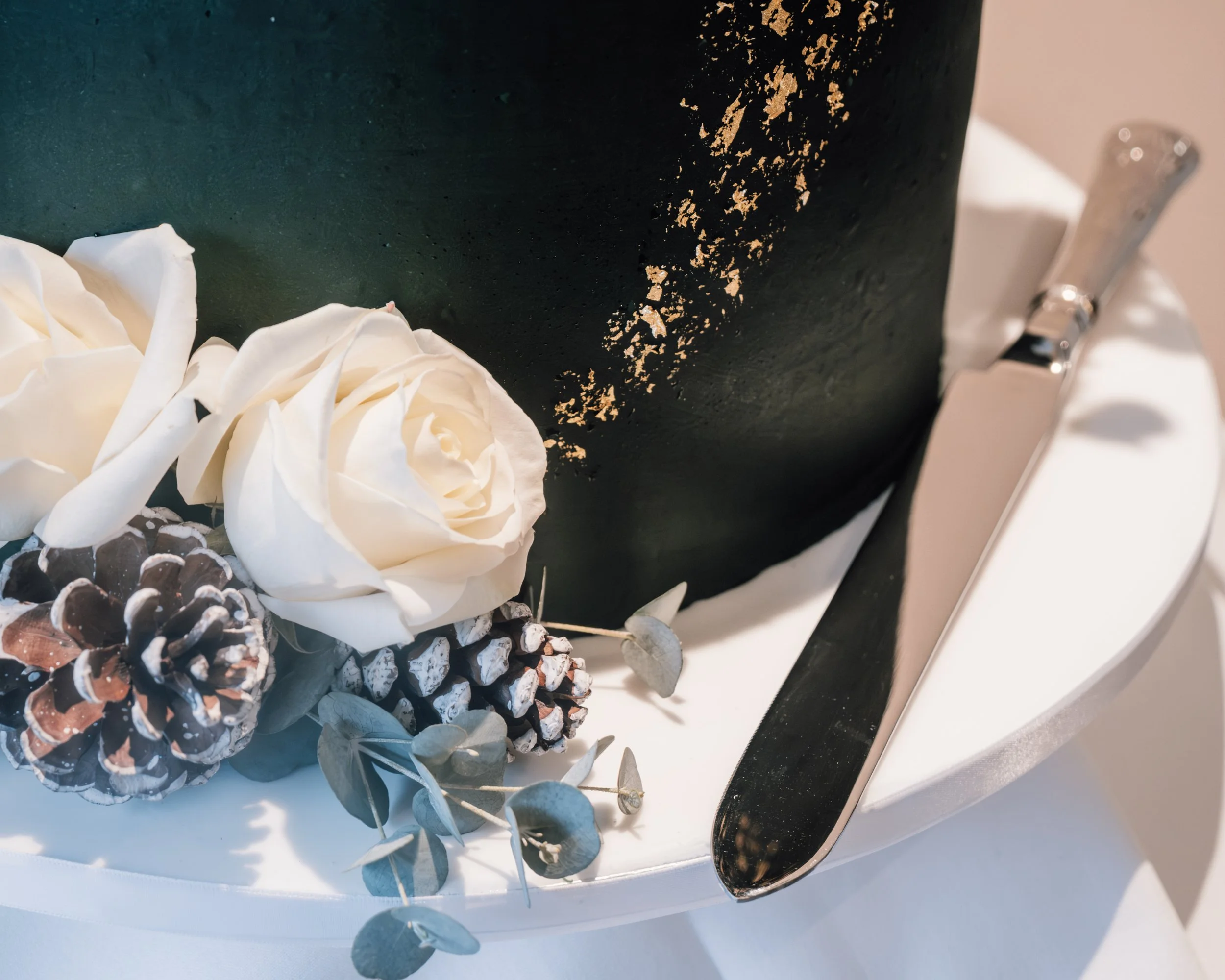 Close-up of a decorated cake with white flower-like icing decorations, pinecones, and eucalyptus leaves, with a dark green textured surface and gold accents on the cake.
