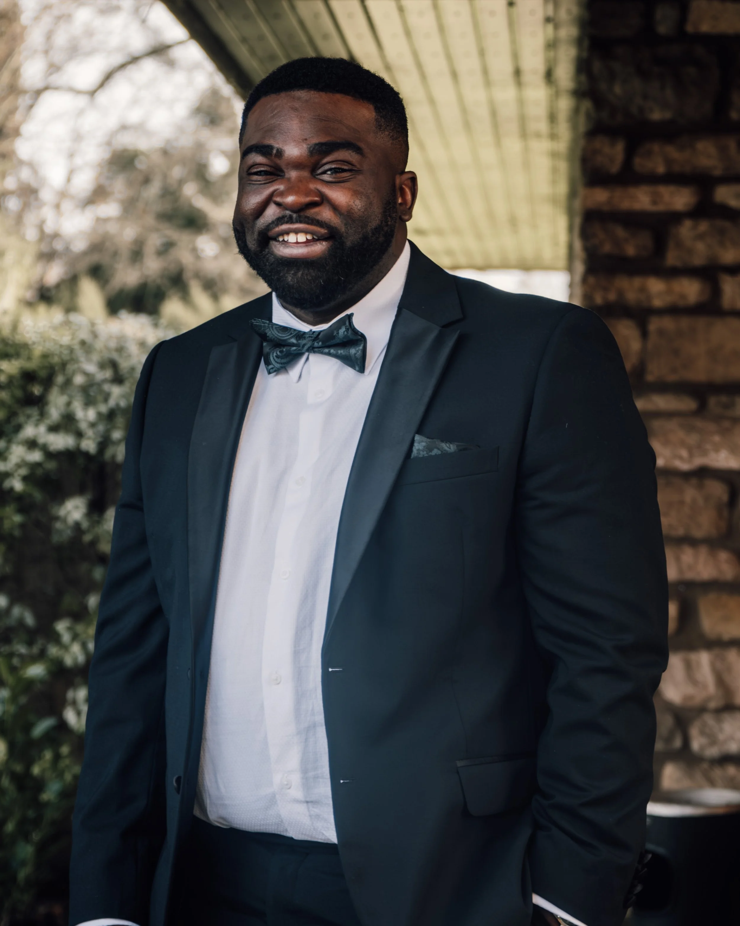 A man wearing a black tuxedo with a white shirt and a patterned black bow tie, smiling outdoors near a stone wall with trees in the background.