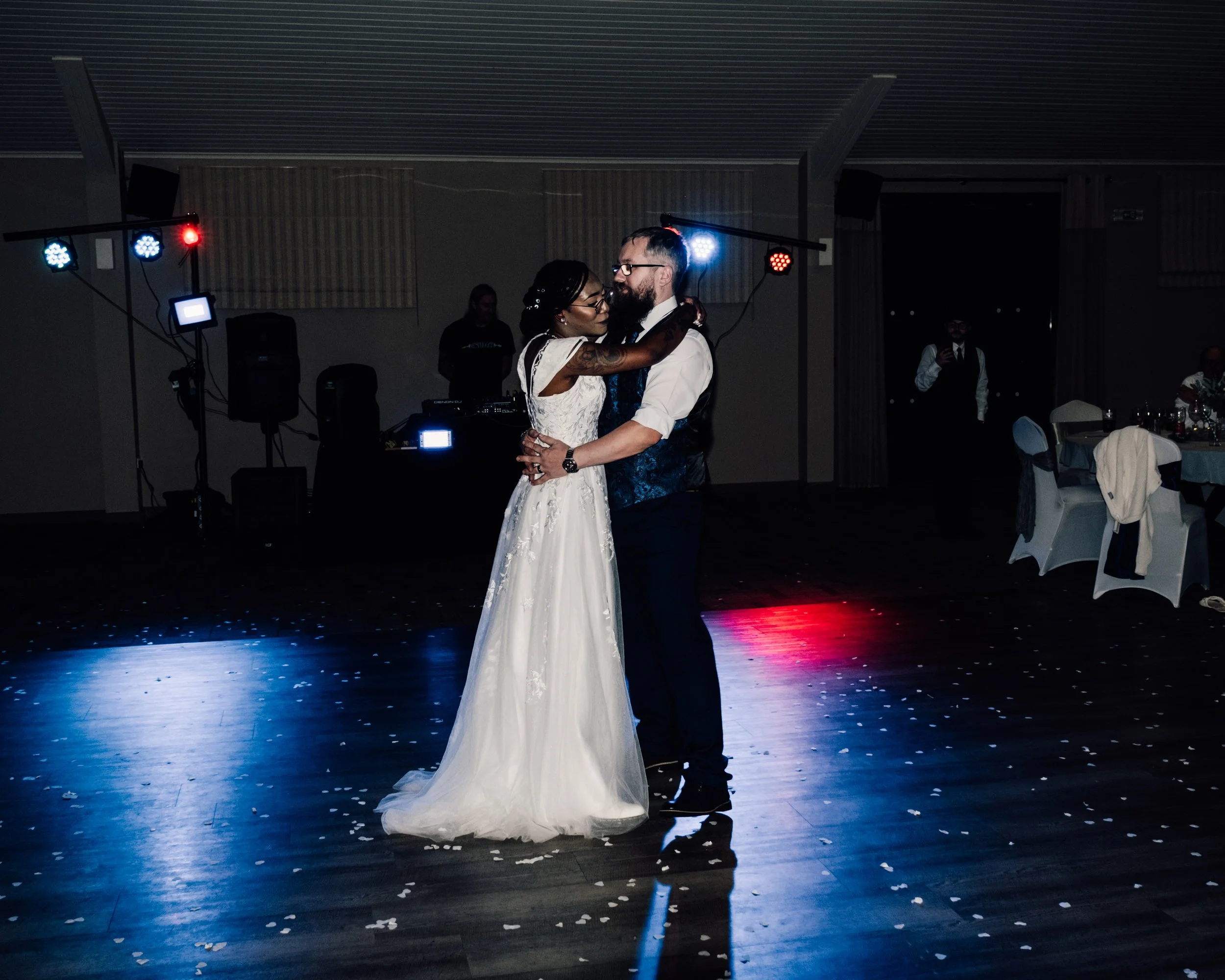 A bride and groom dancing at their wedding reception with dim lighting, colorful stage lights, and scattered flower petals on the floor.