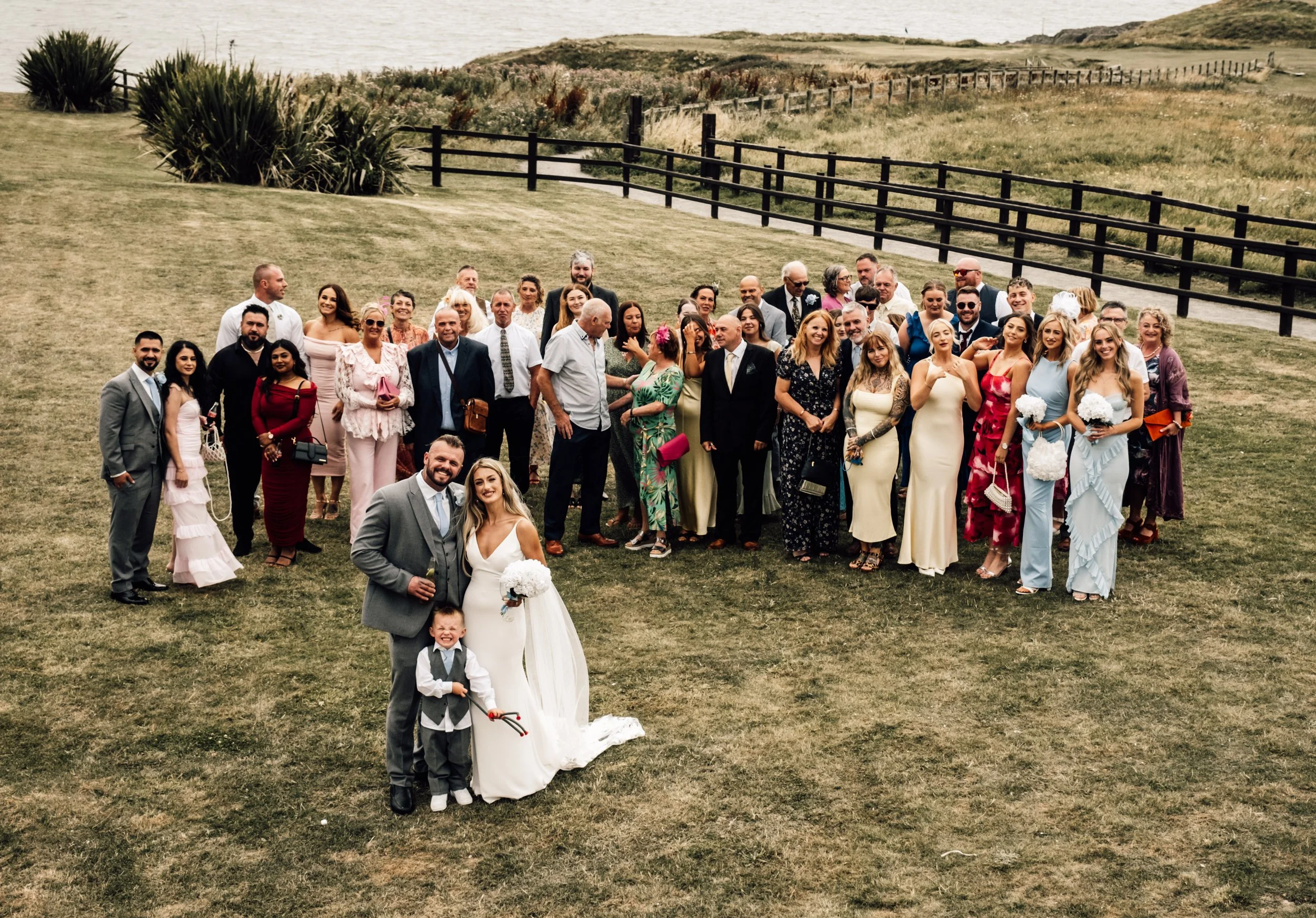 A large group of people dressed in formal attire, celebrating outdoors near the coast on a grassy area with a fence and rolling hills in the background, suggesting a wedding or special event.