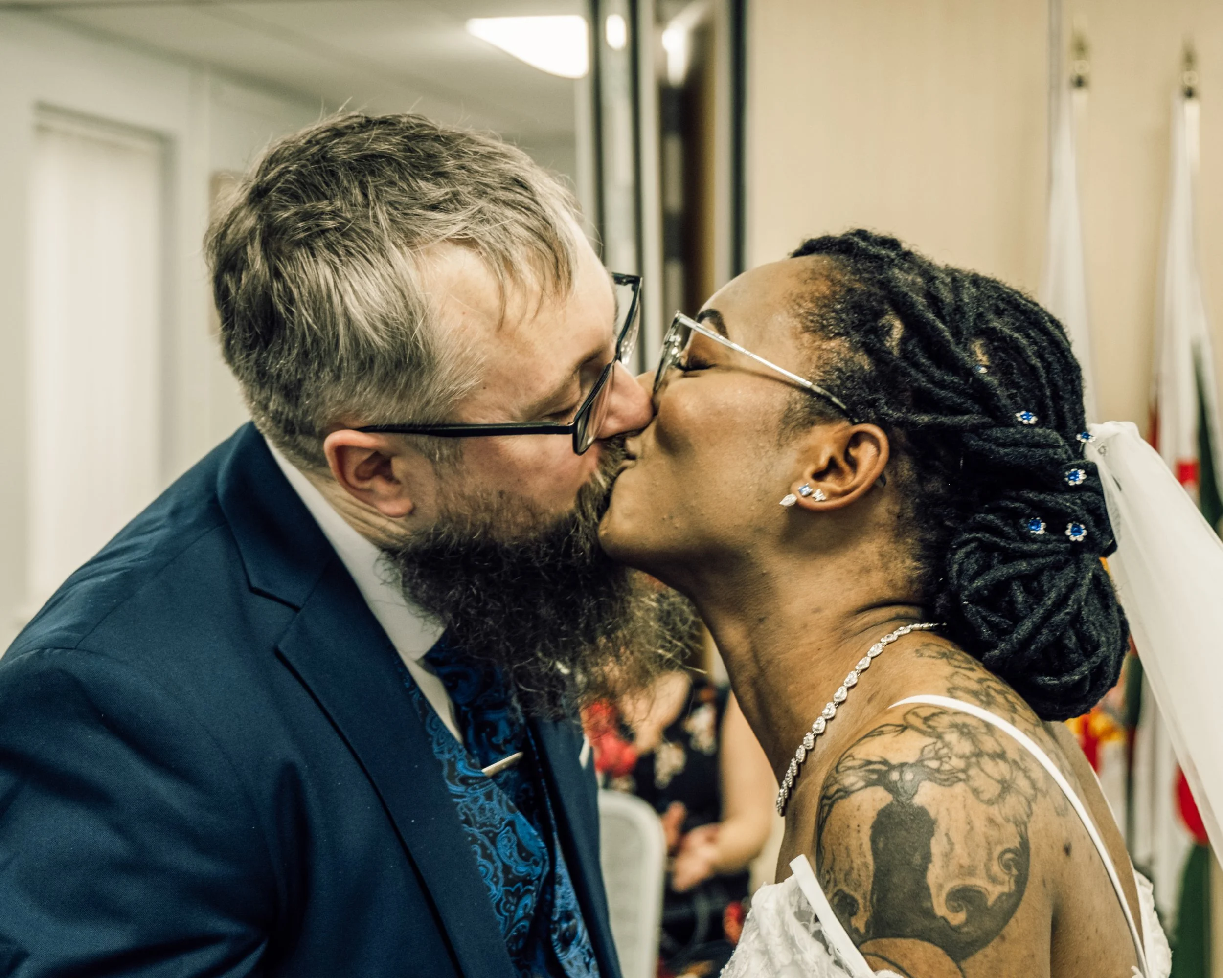 A man in a suit and glasses is kissing a woman with dreadlocks and tattoos on her shoulder, who is wearing earrings and a necklace.
