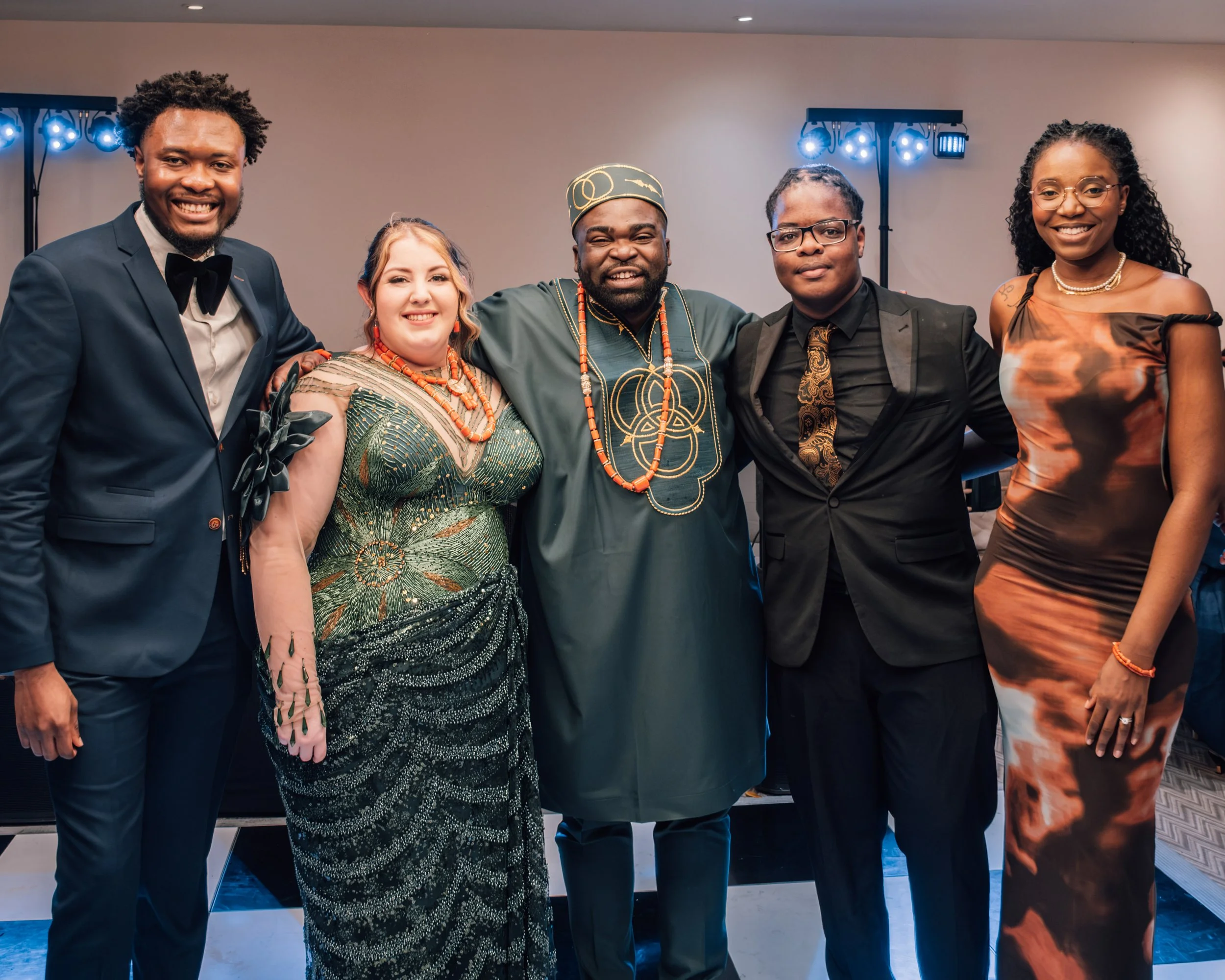 Group of six diverse people dressed in formal and traditional attire, posing together indoors at a celebration or event.