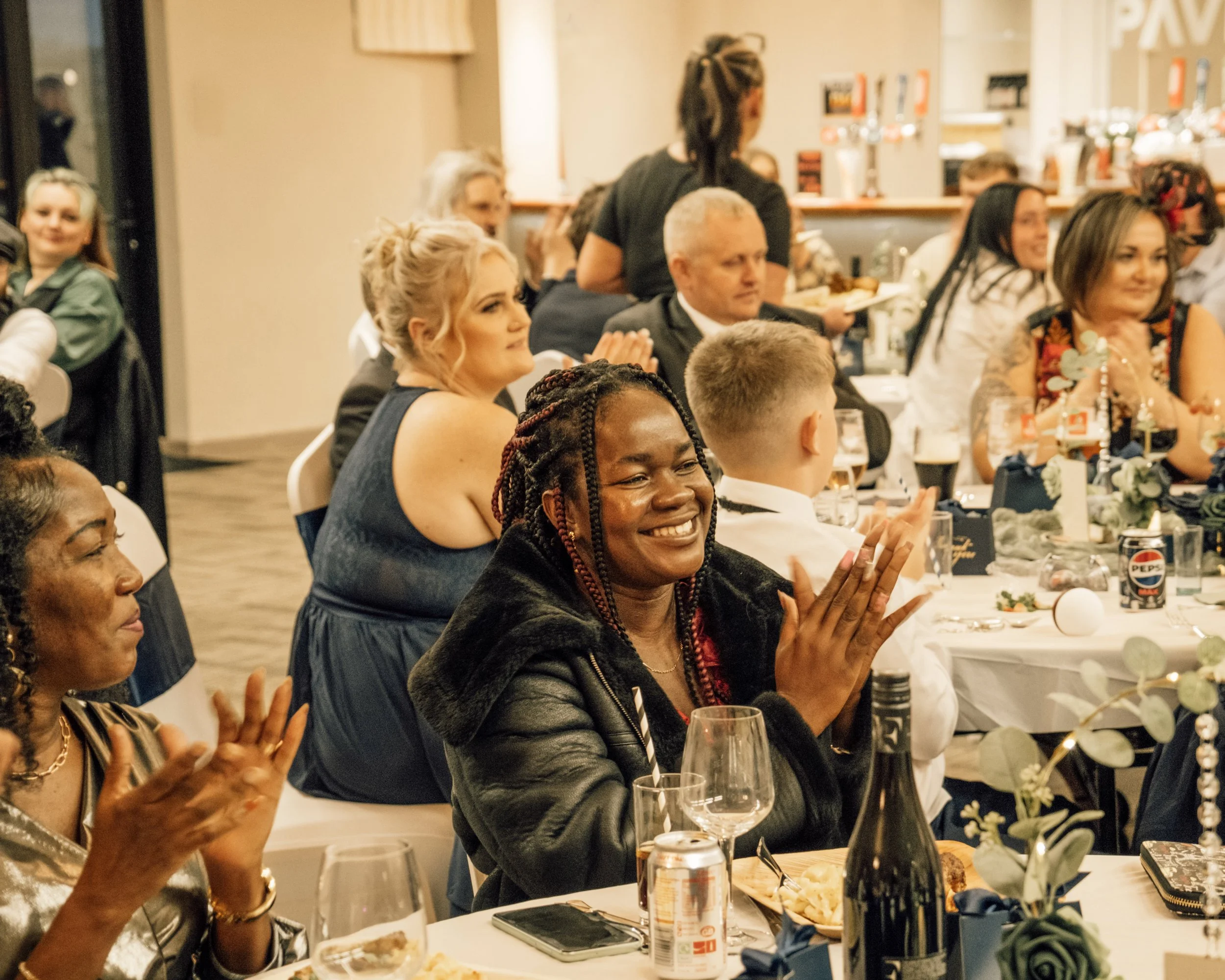 People sitting at a dinner table applauding, with drinks, food, and decorations visible, at a social event or celebration.
