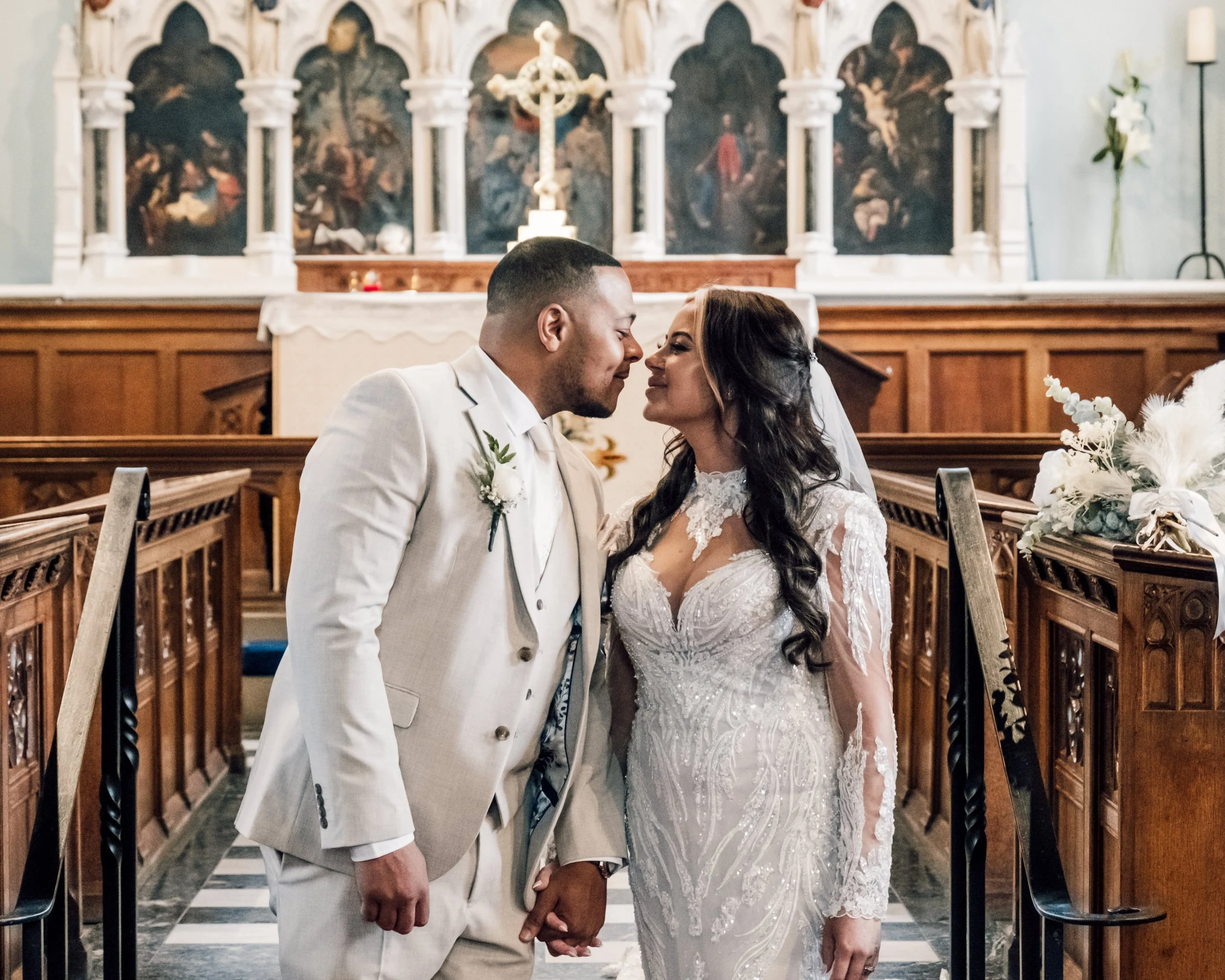 A bride and groom kneeling in a church altar, holding hands and touching foreheads, during their wedding ceremony. The background features religious artwork and a cross.