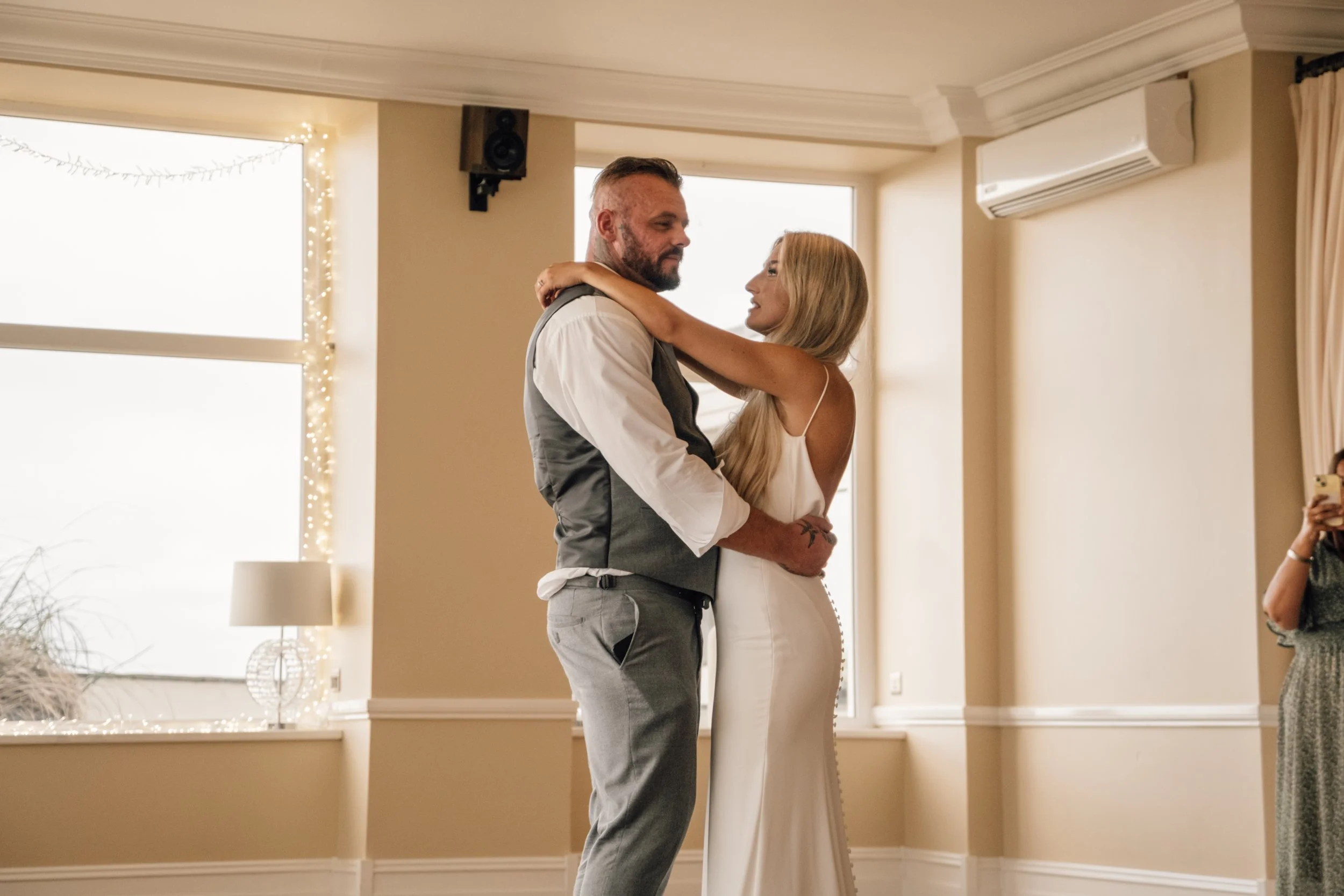 A man and woman dancing closely indoors with natural light, decorated with string lights, in a warmly lit room.
