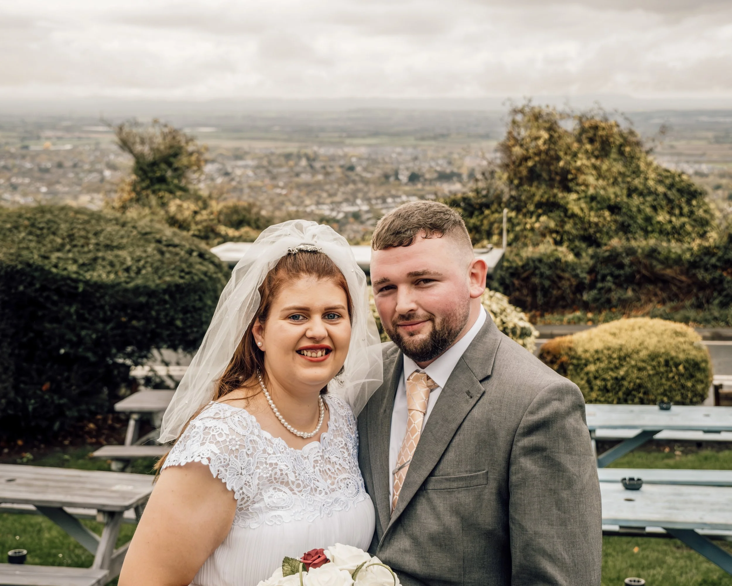 A bride and groom smiling outdoors on their wedding day, with trees and a cityscape in the background.