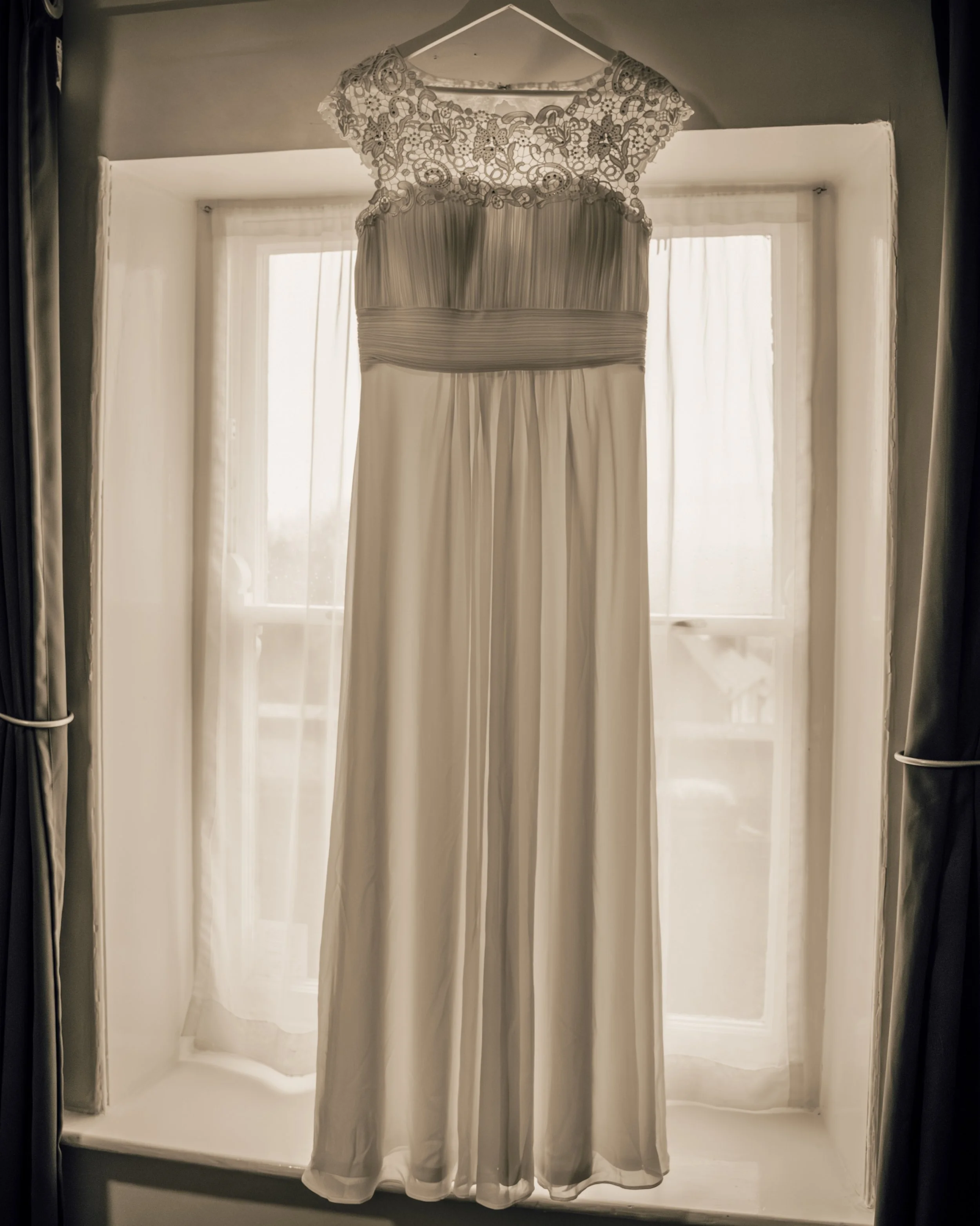 A white wedding dress with lace cap sleeves hanging on a hanger in a window seat with sheer curtains, backlit by sunlight.