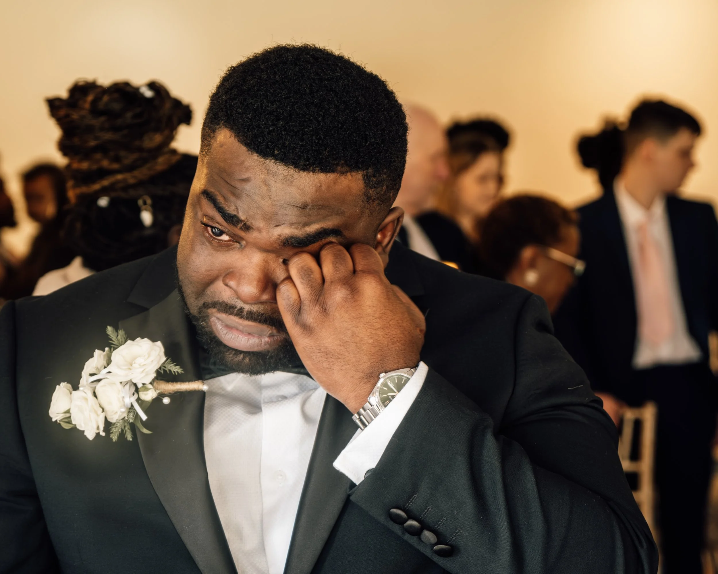 A man in a tuxedo wiping his eye during a wedding ceremony, with a wedding band and a white flower boutonniere on his lapel, surrounded by seated guests.