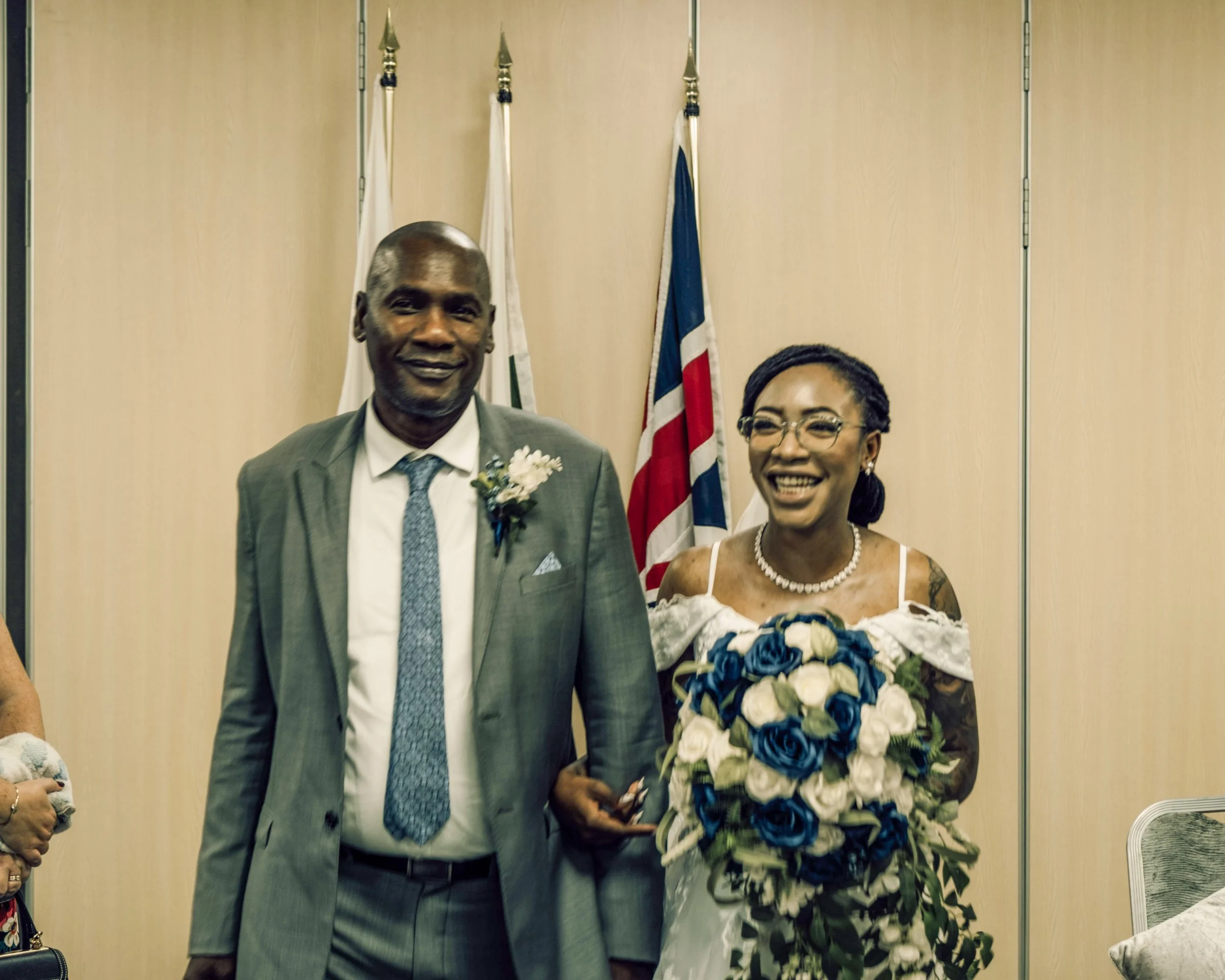 A man and a woman standing together at a formal event, with flags in the background. The man is dressed in a suit with a boutonnière, and the woman is holding a large bouquet of white and blue roses, wearing glasses and a necklace.