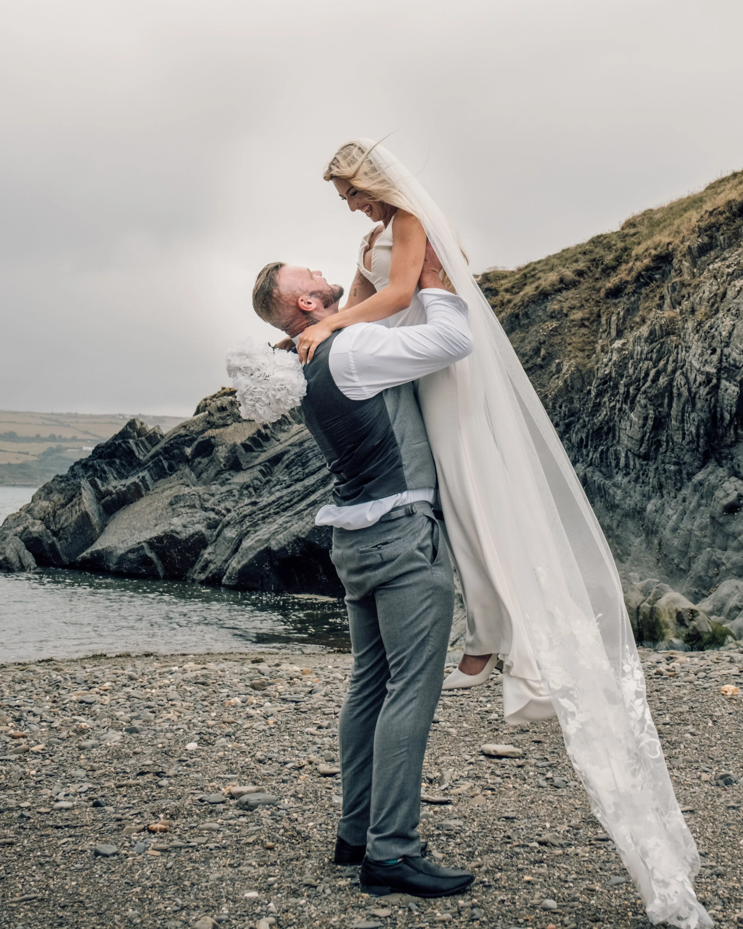 A joyful couple dressed in wedding attire on a rocky beach, with the groom lifting the bride and both smiling, against a cloudy sky and a hillside in the background.