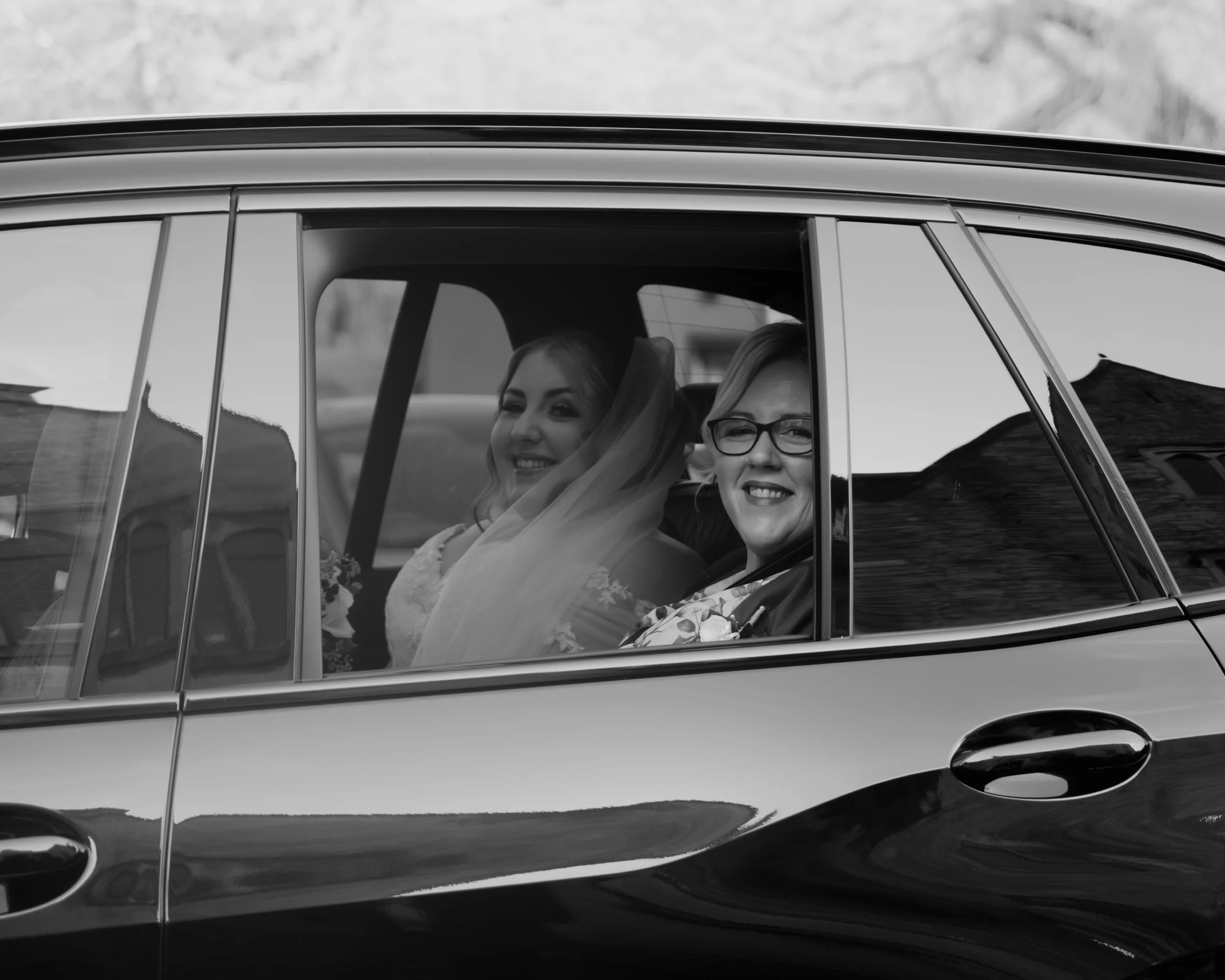 A black and white photograph of two smiling women sitting inside a car. The woman in the front is wearing glasses and has short hair, while the woman in the back has long hair and is smiling.