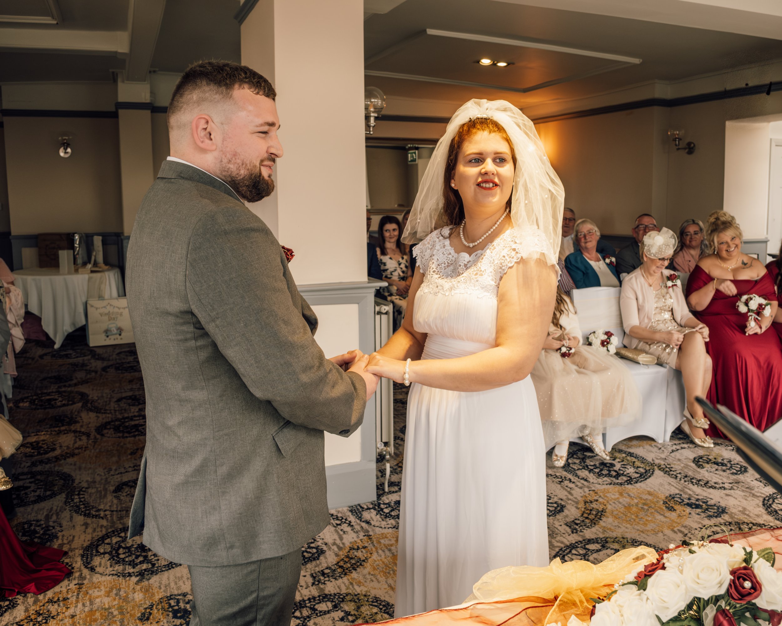 A bride and groom holding hands during their wedding ceremony in a decorated indoor venue, with seated guests watching.