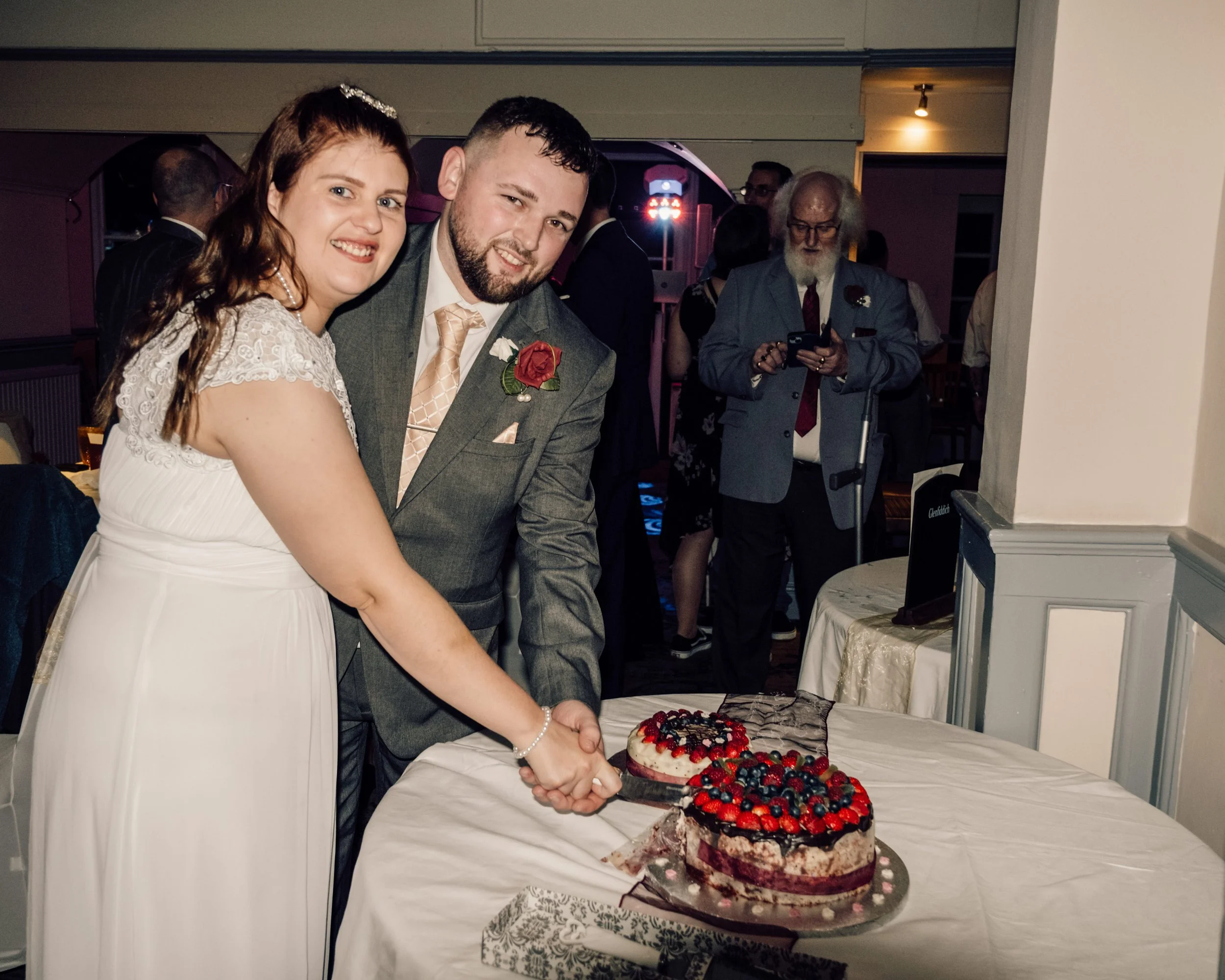 A bride and groom cut a wedding cake decorated with various berries at their wedding reception. The bride has long, red hair and wears a white dress with lace sleeves; the groom has dark, curly hair and a beard, and wears a gray suit with a pink tie 