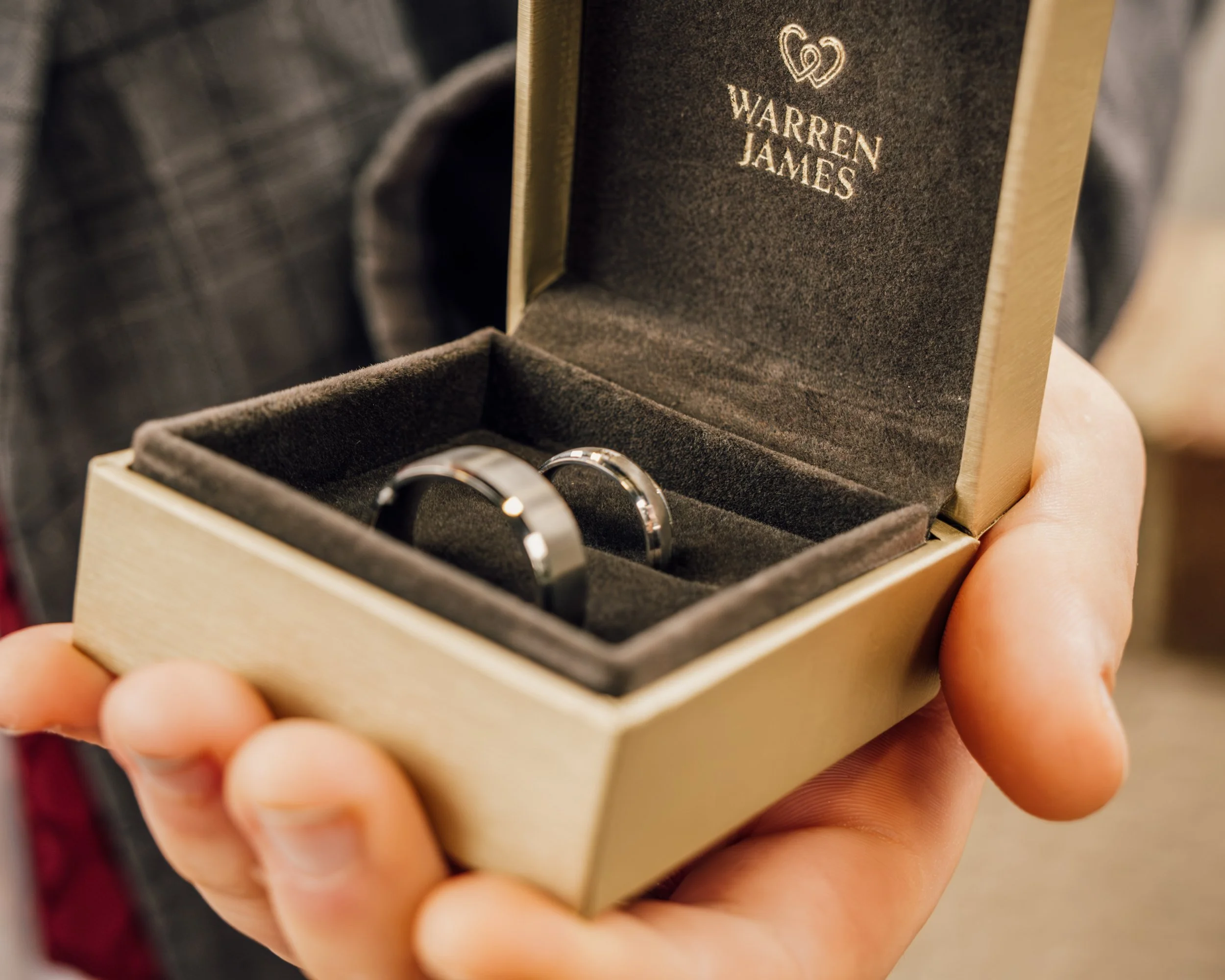 A hand holding a jewelry box with a pair of rings inside, with the name 'Warren James' on the inside of the box lid.