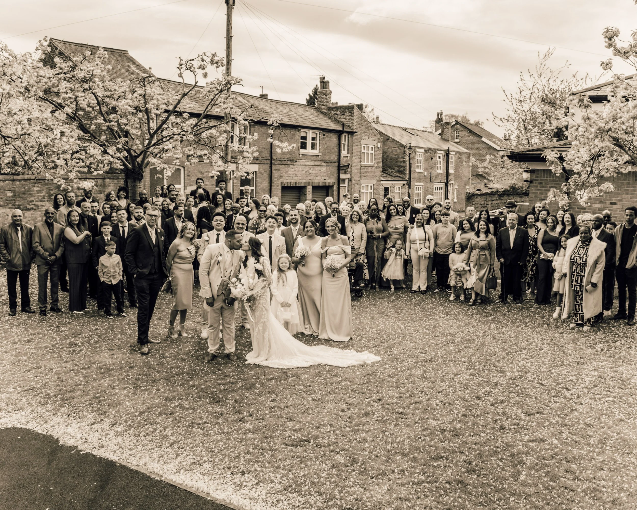 A large group of people gathered outside on a street with houses and blooming trees, dressed in formal attire, for a celebration or event.
