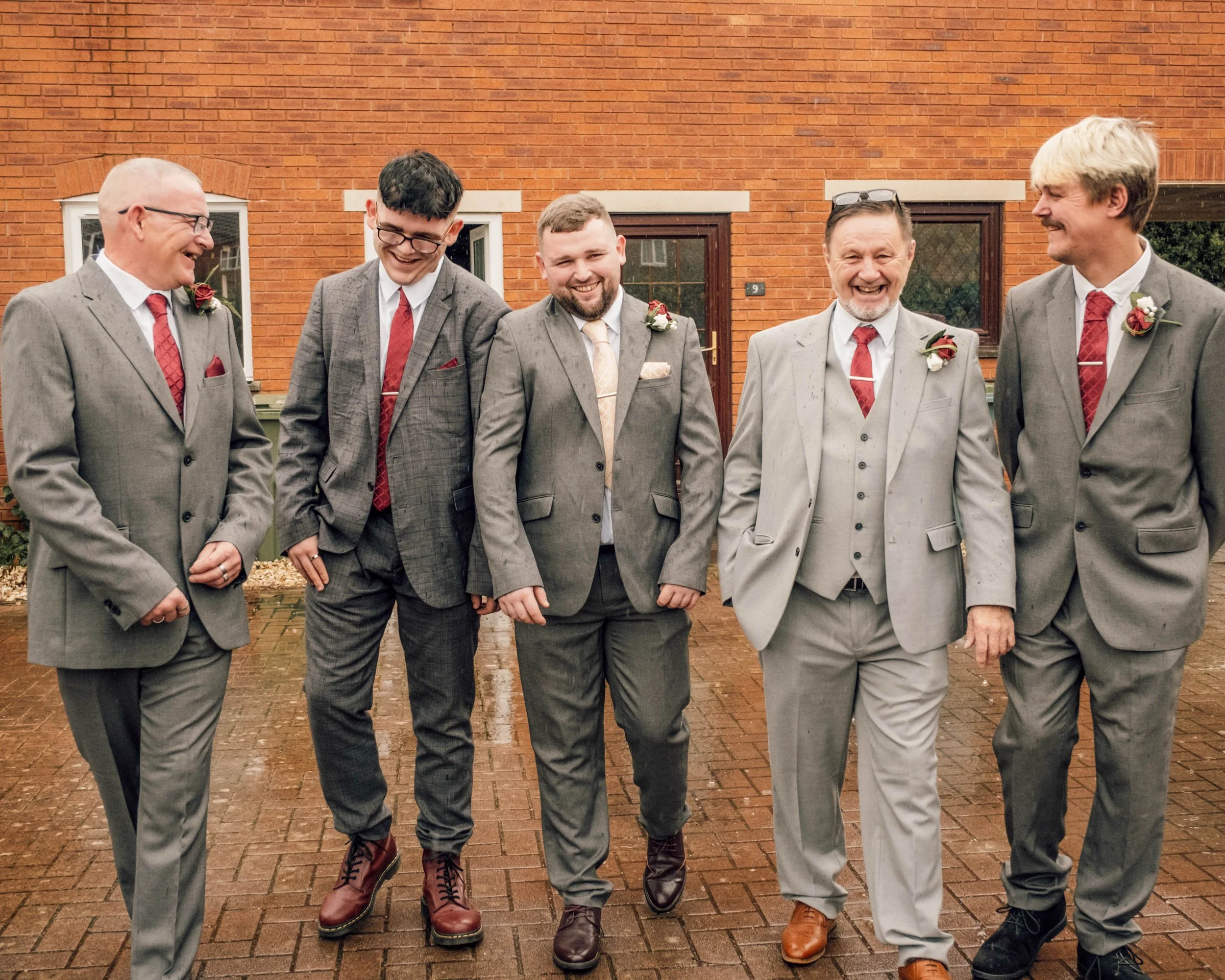 Group of five men dressed in suits standing outside in front of a brick wall, smiling and engaging with each other.