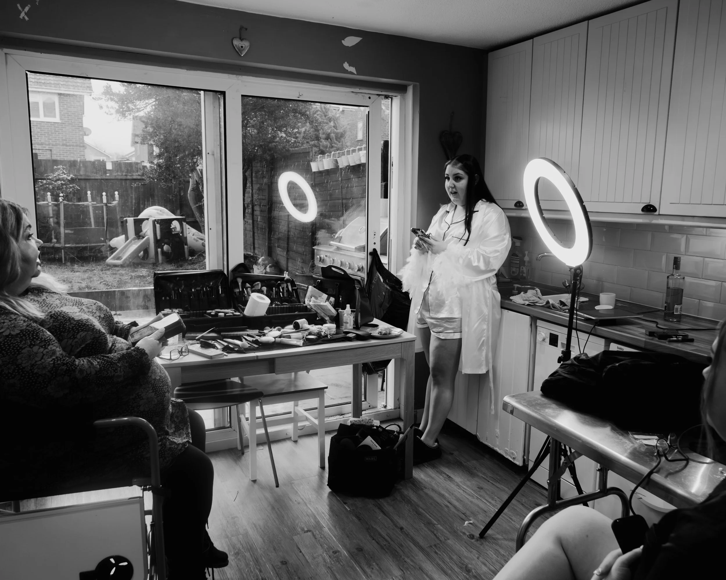 A woman wearing a white coat is standing in a kitchen, using her phone. Two people are seated at a table with makeup tools and brushes. A ring light and a large window showing a backyard are also visible.