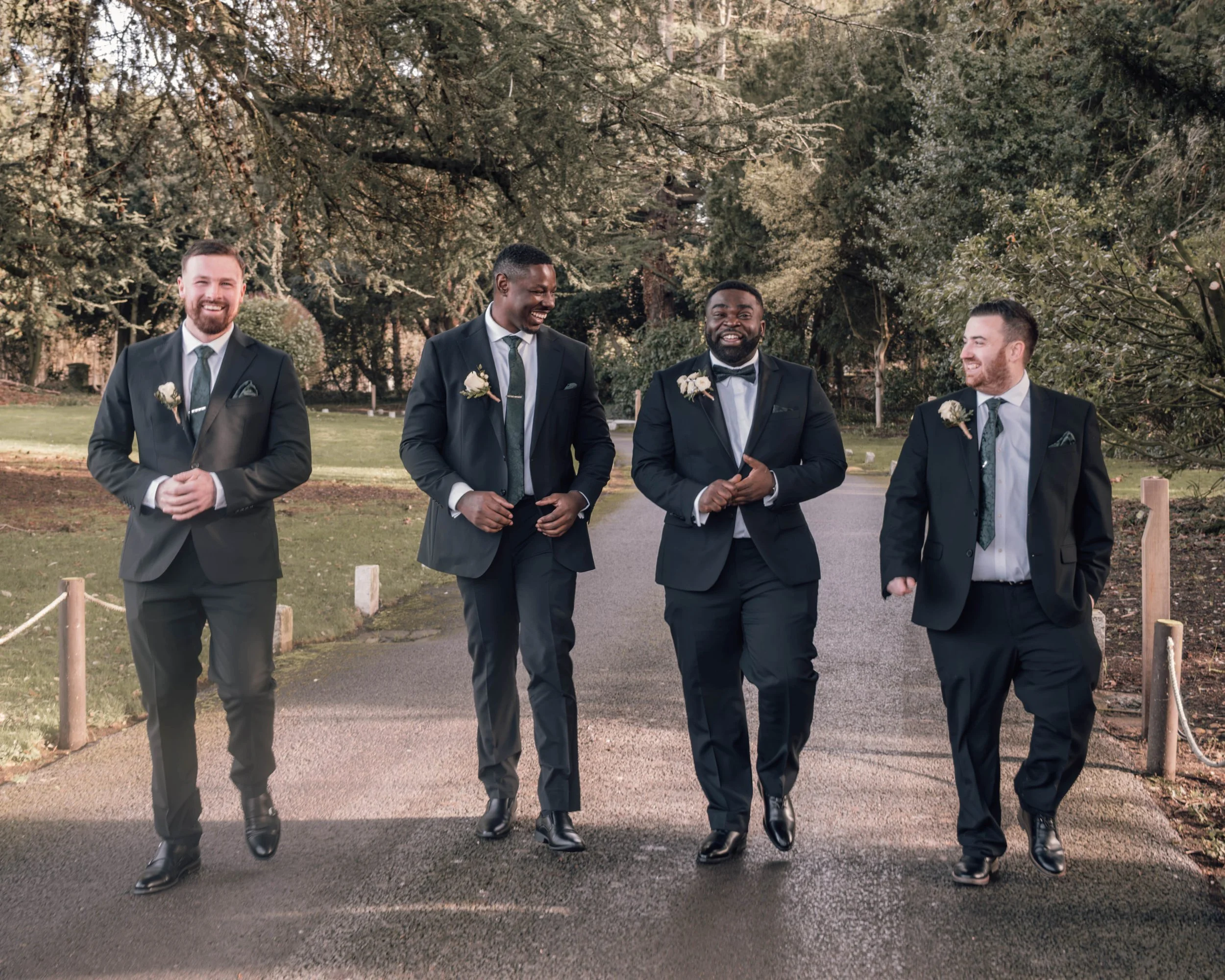 Four men in suits walking outdoors in a park-like setting. The trees behind them are large and leafy, and the men are smiling and chatting as they walk.