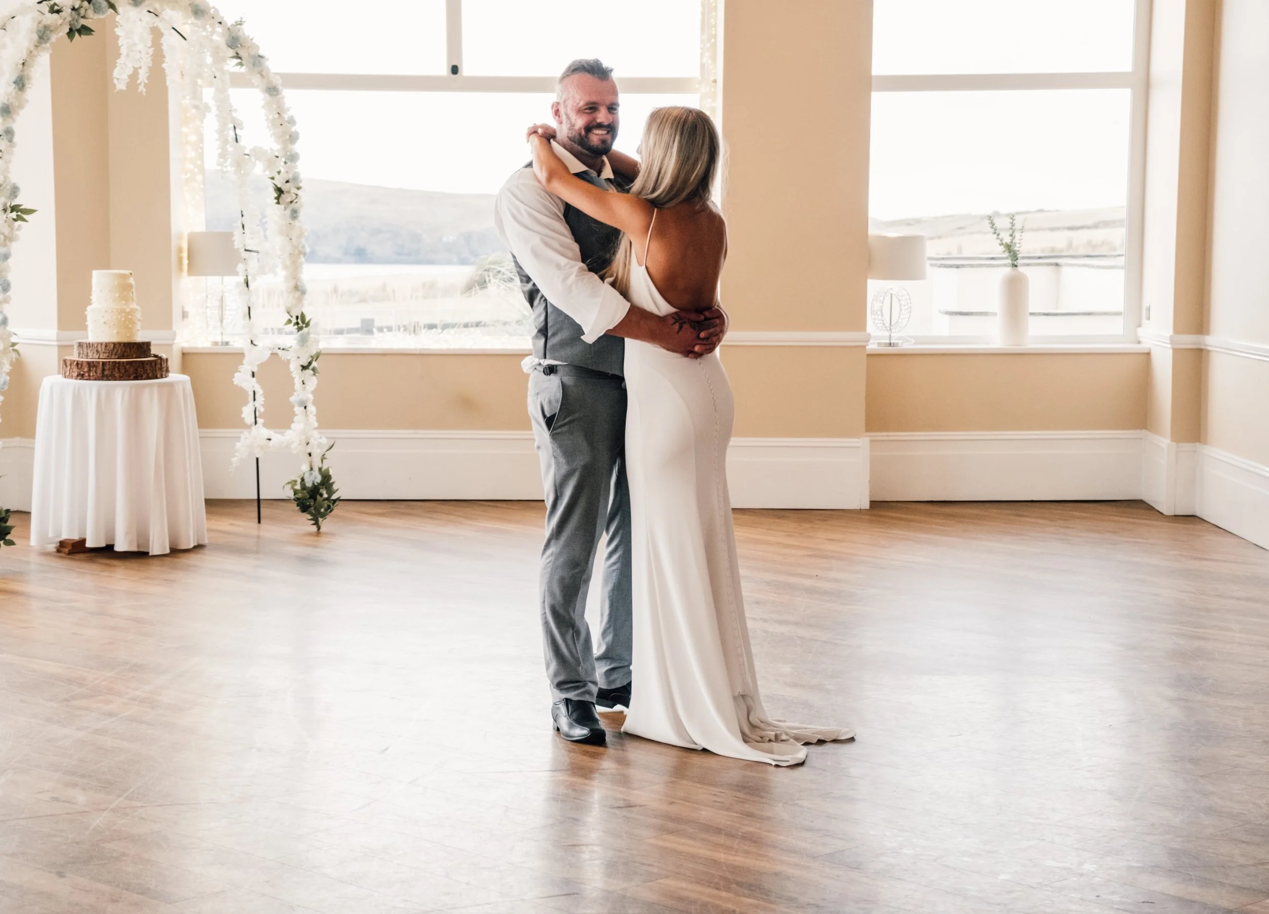 A bride and groom sharing their first dance at a wedding reception in a room with large windows and minimal decorations.