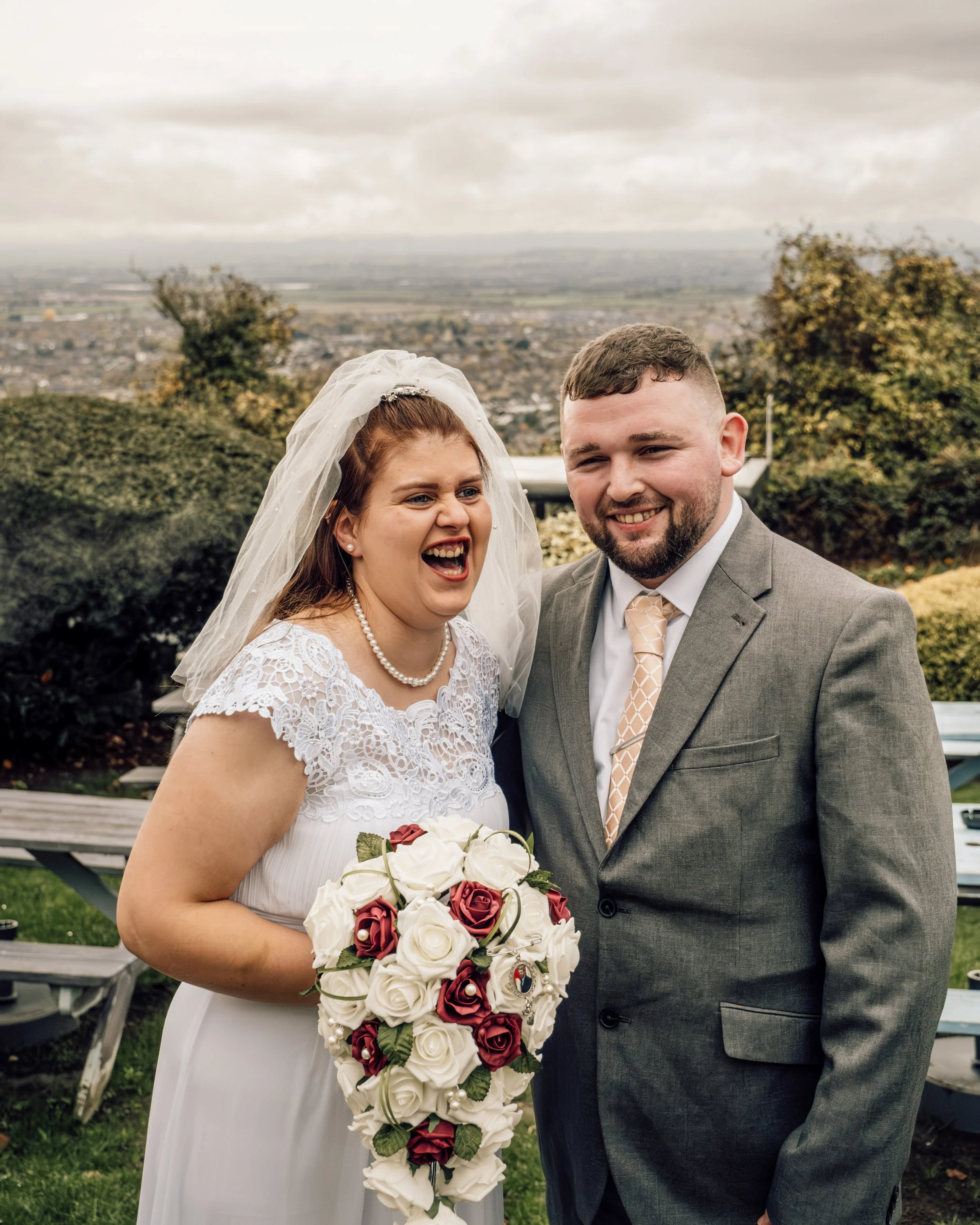 A bride and groom smiling outdoors, holding a bouquet of white and red roses, with a view of a cityscape and cloudy sky in the background.