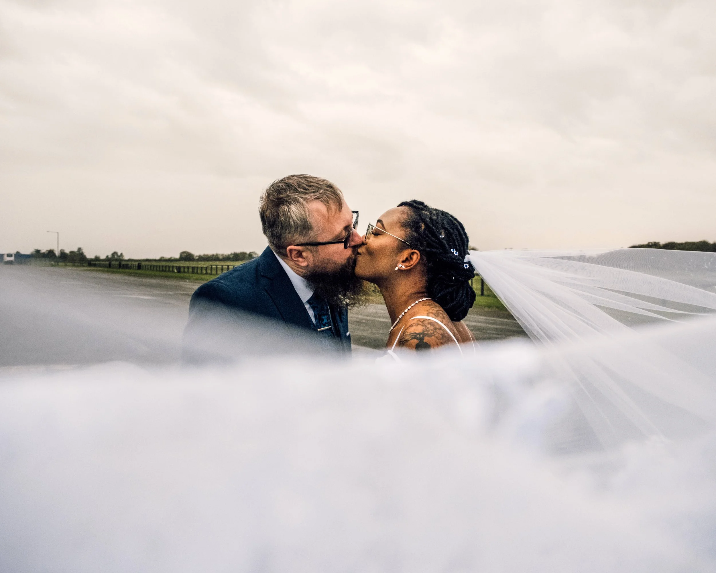 A couple sharing a kiss outside on a cloudy day, with a landscape of green fields and a wooden fence in the background. The woman has braided hair and tattoos, and the man has glasses and light hair. The bride's veil is flowing.