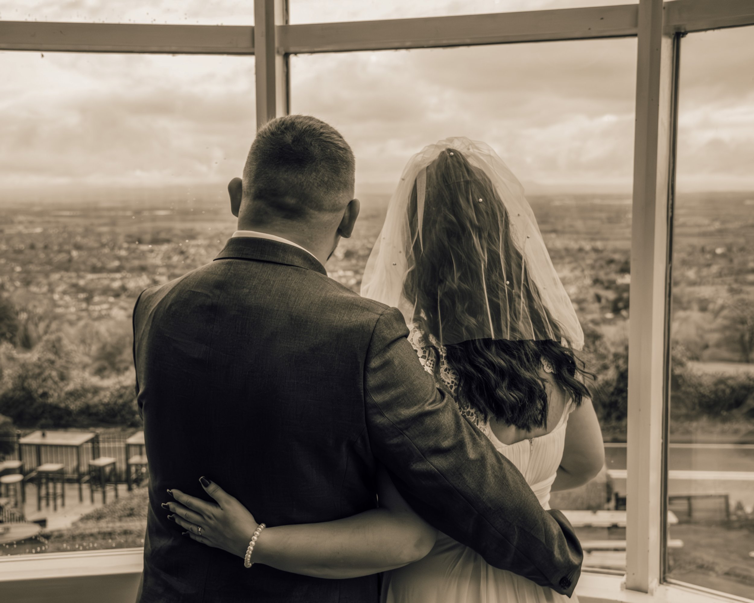 A couple dressed in formal attire, with the man wearing a suit and the woman wearing a wedding dress and veil, sharing a romantic moment as they gaze out a window at a cloudy sky and a landscape.