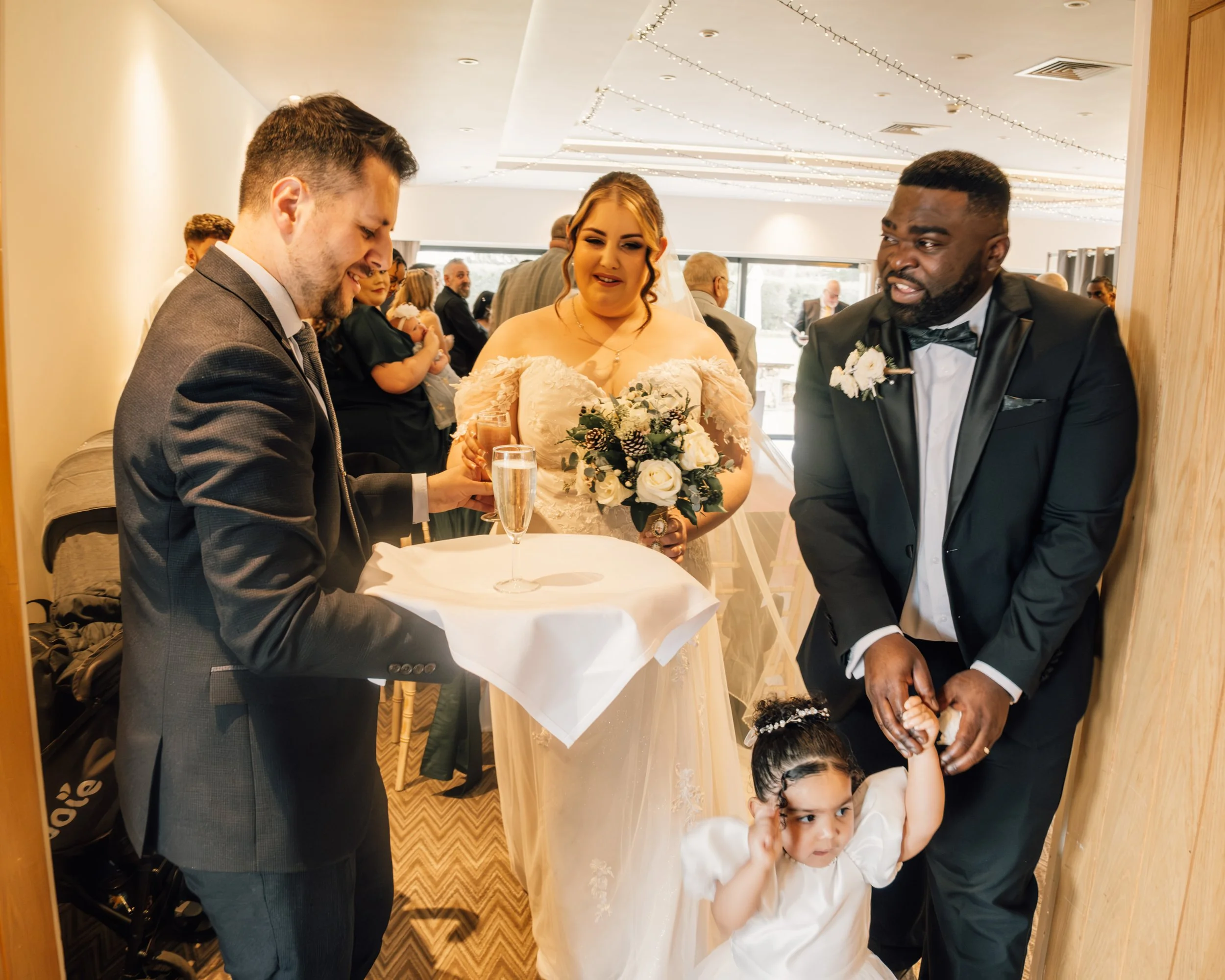 A bride in a wedding dress holding a bouquet, a man in a tuxedo, and another man in a suit sharing a toast at a wedding reception, with guests in the background.