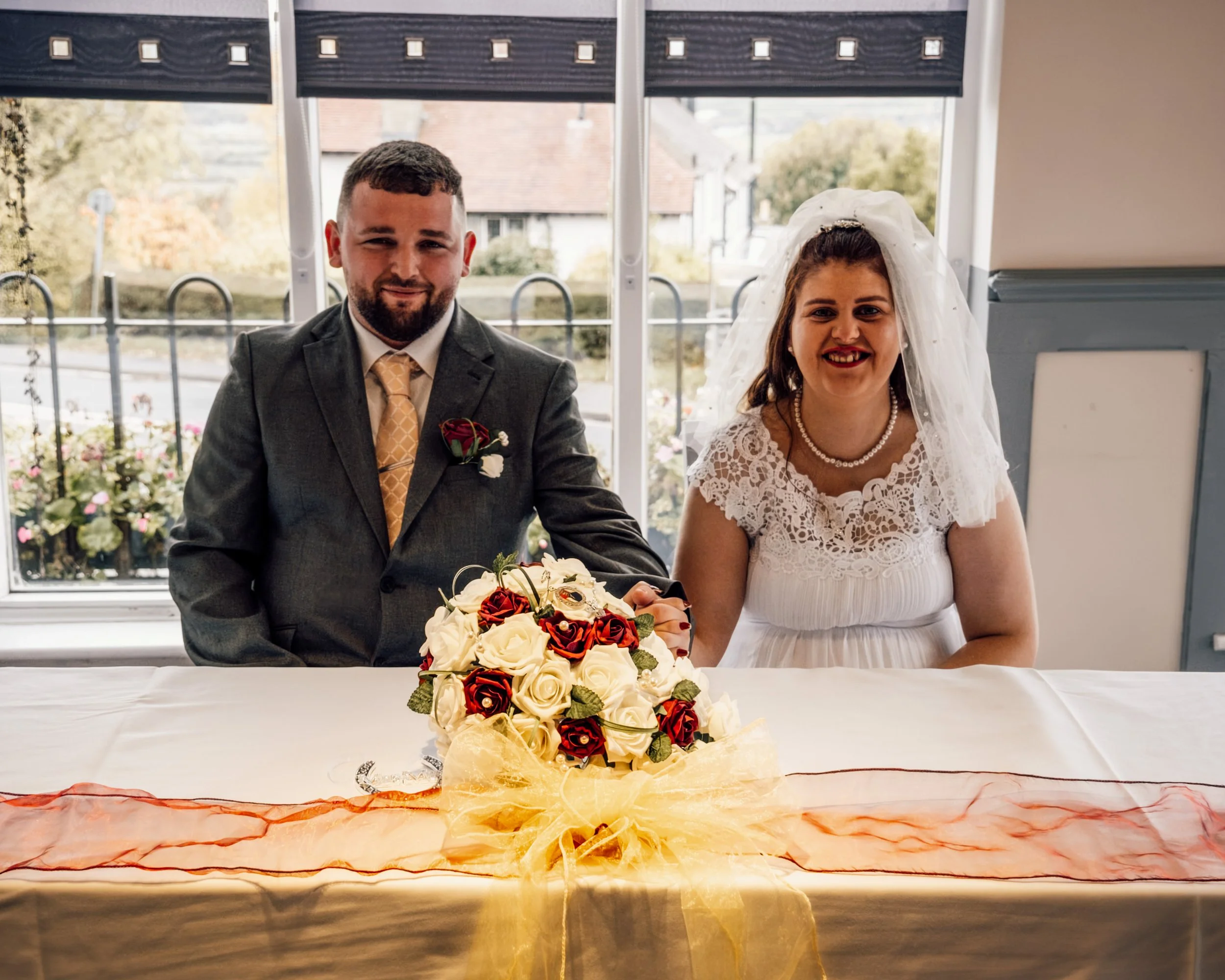A man and woman dressed in wedding attire sitting at a table with a floral cake in front of them. The man is wearing a suit and tie, and the woman is wearing a white lace dress and veil.